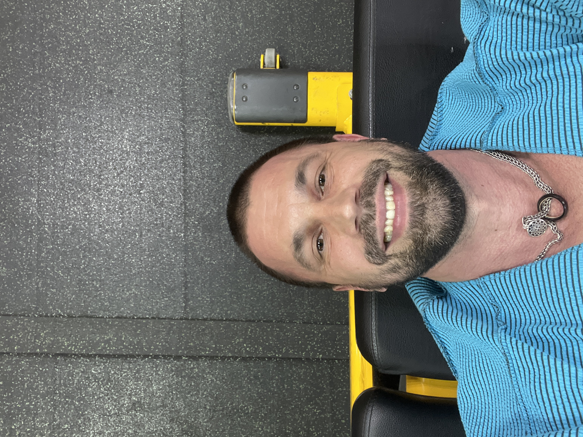 A man with a wide, bright smile is captured in a close-up, likely a selfie, within what appears to be a gym setting. He has short dark hair, a neatly trimmed beard, and a mustache. His head is slightly tilted, and his eyes are looking directly at the viewer. He is wearing a light blue shirt with thin black vertical stripes and a silver chain necklace with a decorative pendant featuring a ring and a multi-pointed star design.
The man is positioned against black padded upholstery, which is part of a piece of gym equipment, possibly a bench or a machine. A yellow frame is visible behind and to the side of him, supporting the black padding. To the left of his head, a gray and yellow mechanical component, possibly part of the adjustment mechanism for the gym equipment, is visible, featuring a small wheel on top. The background is a dark gray or black speckled floor, characteristic of rubber gym mats, with faint lines suggesting seams between mat sections. The overall impression is one of happiness or contentment, given the man's broad smile. There is no visible text in the image.
