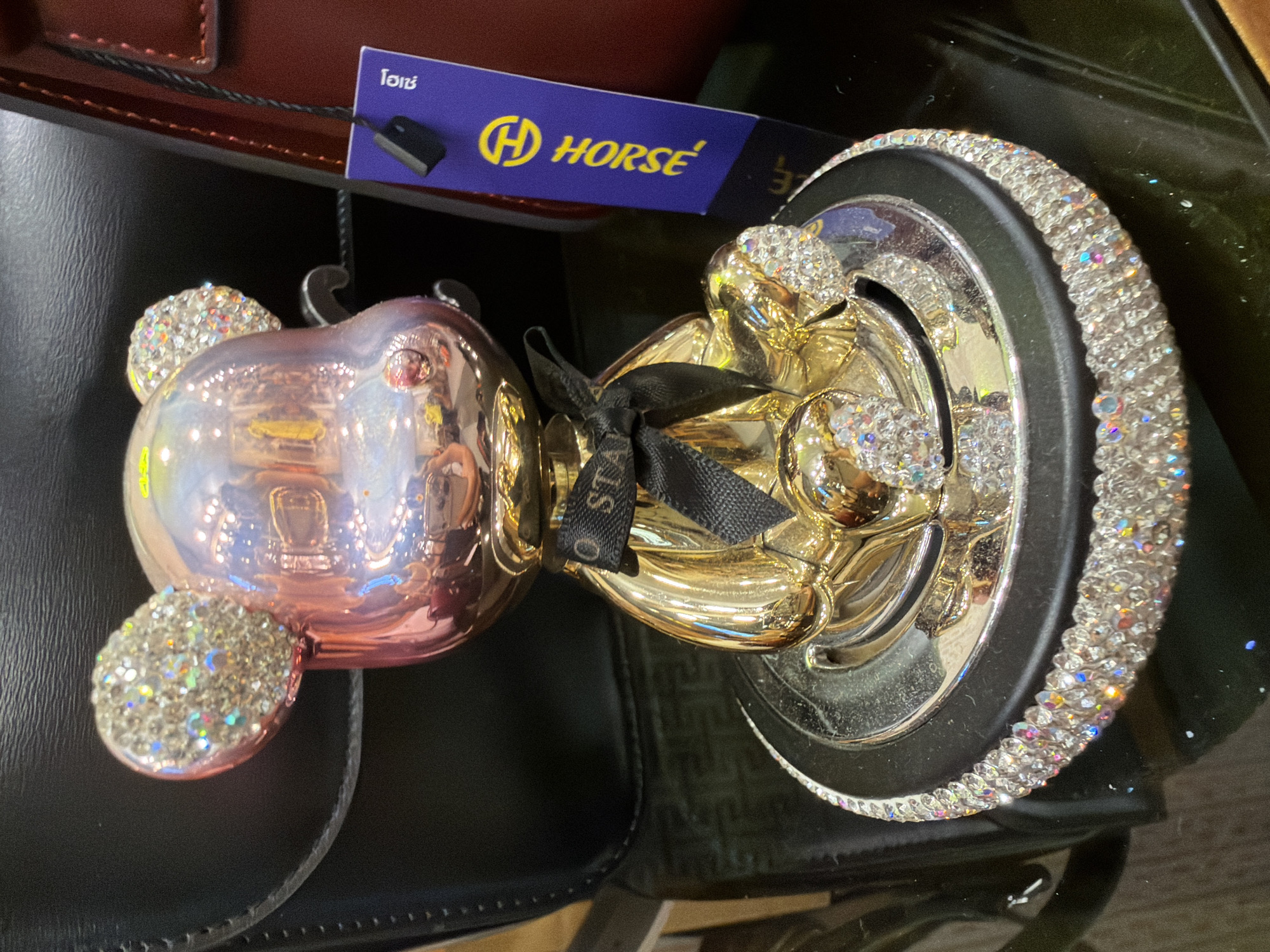 This media file features a close-up, angled shot of a decorative object, possibly an ornament or a car accessory, placed on a dark, reflective surface, likely a shelf or tabletop. The object itself is a stylized teddy bear head with a rose-gold metallic finish. It is adorned with numerous small, clear crystals, particularly on its ears and face, which catch the light and create a dazzling effect.
The teddy bear's head is attached to a golden base, also encrusted with crystals. A black ribbon is tied around the "neck" area of the teddy bear. The base has a black rim that is also heavily decorated with the same clear crystals. The overall impression is one of luxury and opulence.
In the background, a purple tag is visible, attached to a dark cord. The tag has a yellow logo featuring a stylized "H" inside a circle, followed by the word "HORSE" in yellow lettering. There is also some text in Thai script above the logo. The tag appears to be a product label or brand tag. Behind the tag and the main object, glimpses of a dark brown, possibly leather, surface can be seen.
The lighting suggests an indoor setting, possibly a retail environment, given the product tag. The reflective surfaces create interesting light refractions. No people are visible in the frame. The object is the sole focus. The location context provided is Ko Samui District, Thailand, but there are no specific visual cues within the image to confirm this location.