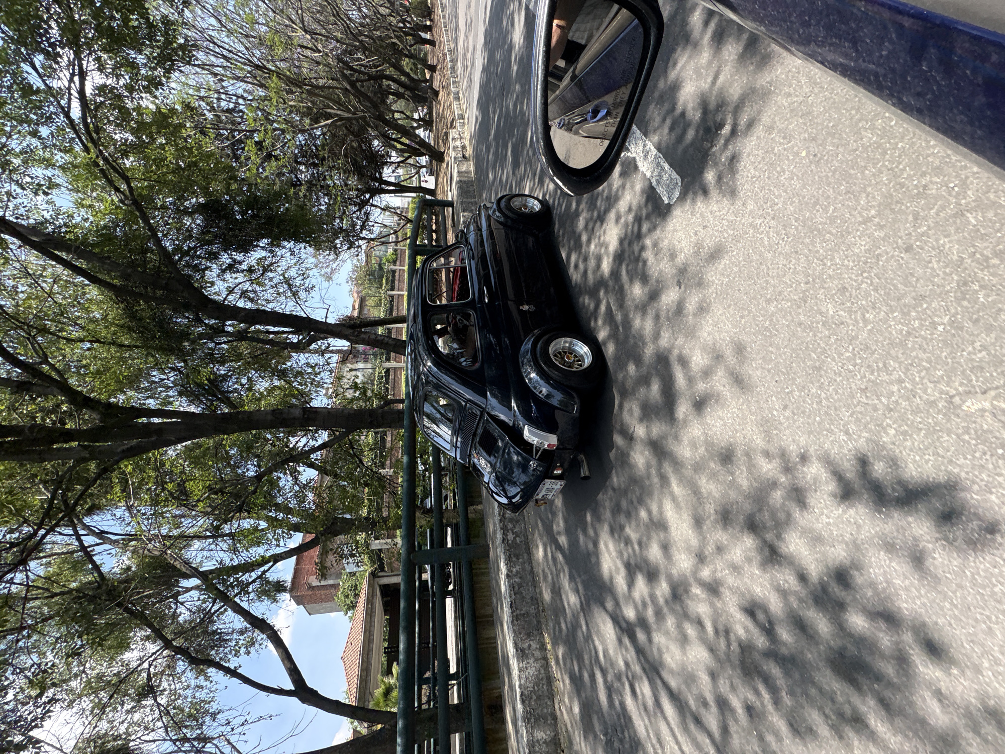 The image shows a black vintage car parked on the side of a road in Mexico City, Mexico. The car is positioned next to a green metal fence, with trees lining the street and casting shadows on the road. A building with a red tile roof is visible behind the fence and trees. A side mirror of another car is visible in the top right corner of the image. The image suggests a sunny day with clear skies.