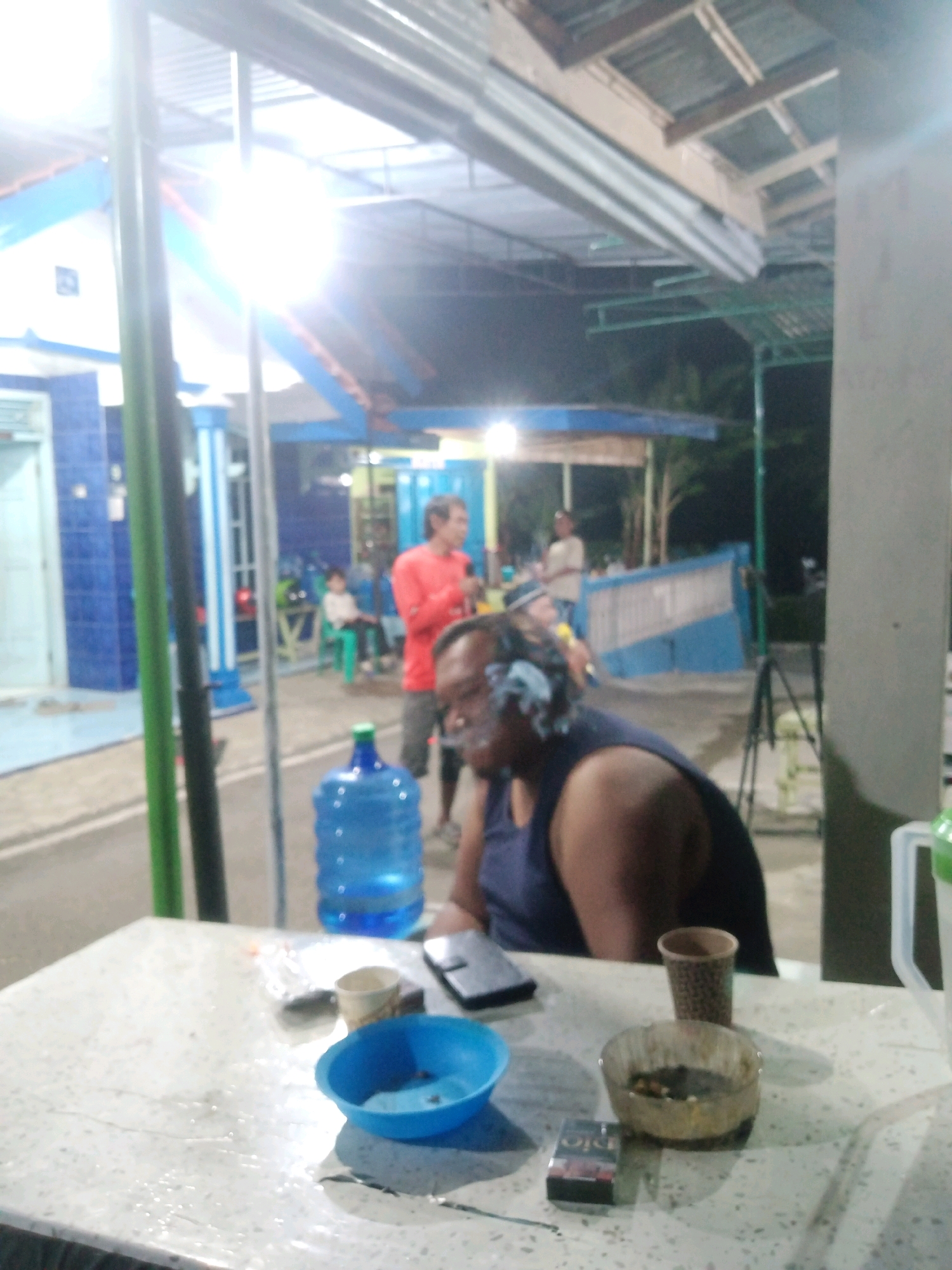 In Rejasari, Indonesia, a person wearing a dark tank top sits at a table, appearing to be smoking. The table holds a pack of cigarettes, an ashtray with cigarette butts, a small blue bowl, a paper cup, and a smartphone. In the background, a large blue water jug is visible. To the left, a building with blue tiled walls and white trim is present. Further in the background, several other individuals are seen, some sitting at tables or standing. The scene appears to be an outdoor seating area, possibly a warung or small eatery, with a covered roof overhead. The lighting suggests it is evening or night.