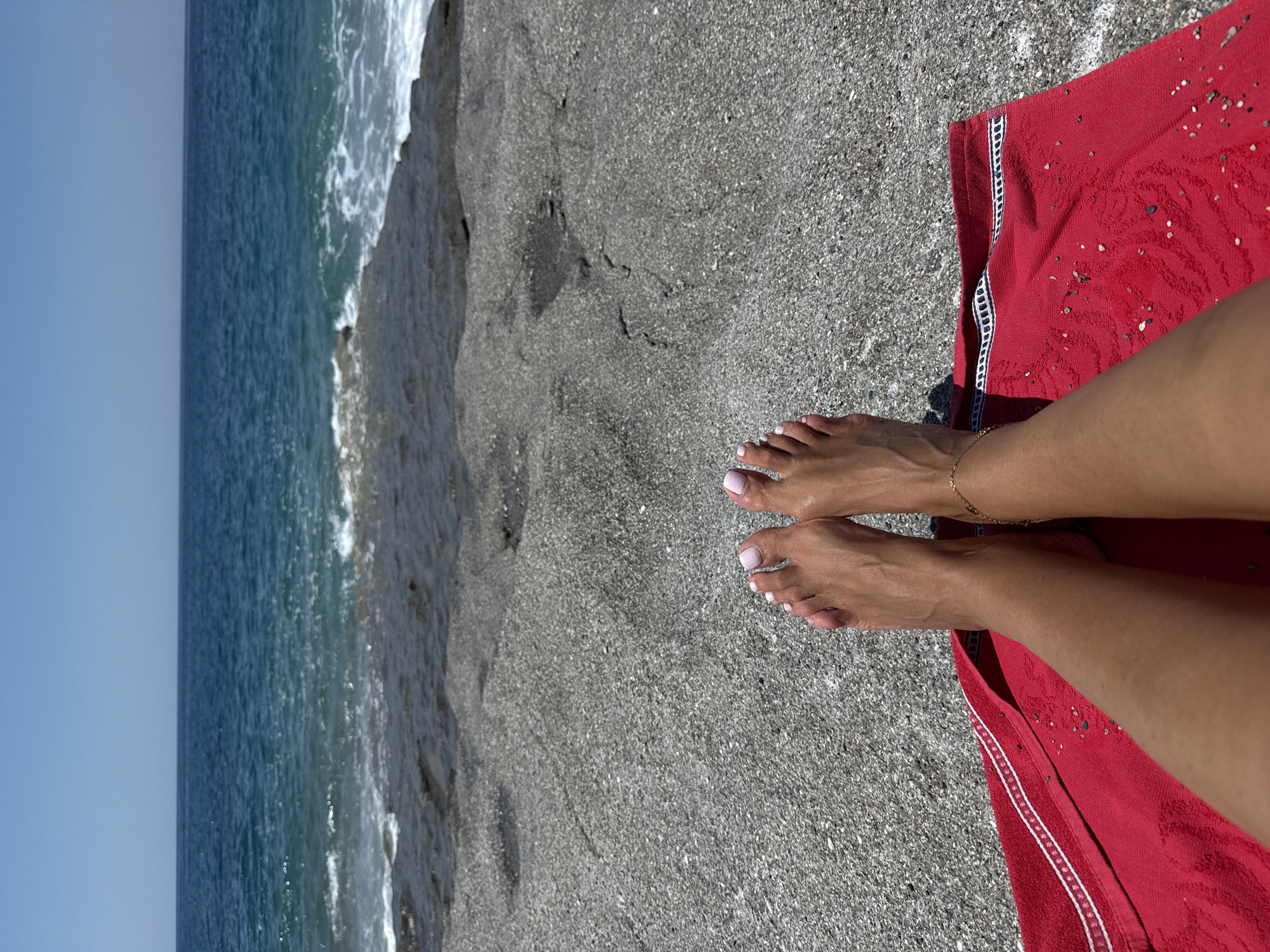 A person's bare feet, with polished light pink toenails and a delicate gold ankle bracelet, rest on a bright red towel spread across a pebbly beach. The towel is positioned in the foreground, partially obscuring the person's legs. Beyond the towel and feet, the beach extends to a darker, rocky shoreline that meets the turquoise Mediterranean Sea. Gentle waves crash against the shore under a clear, bright blue sky. The scene depicts a serene moment on a beach in Alanya, Turkey.