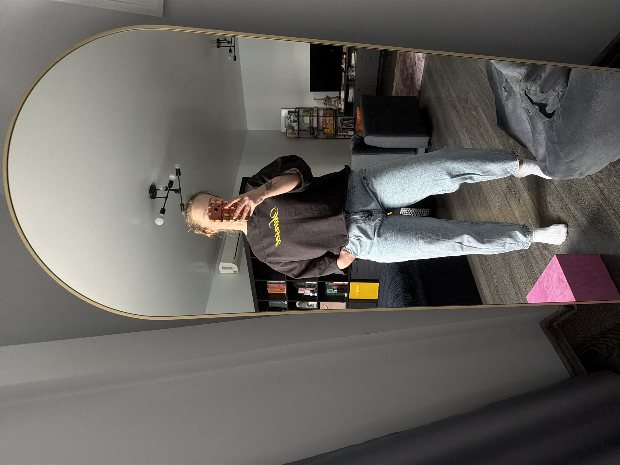 A female individual is depicted in a mirror selfie, captured from approximately mid-thigh up. She has light-colored hair pulled back and is holding a reflective, possibly rose gold, smartphone with a textured case in her right hand, obscuring part of her face. Her left hand is placed in the pocket of her light blue, loose-fitting jeans. She wears a dark, oversized, long-sleeved top with yellow cursive text across the chest and white crew socks.
The mirror is full-length, featuring an arched top and a thin gold-colored frame. The reflection reveals an interior room with light grey walls, dark wooden flooring, and a white ceiling with a modern black light fixture containing multiple exposed bulbs.
Within the reflection, to the left, a dark modular bookshelf is filled with books and various objects, including a prominent yellow box-like item on a lower shelf. An air conditioning unit is mounted high on the wall above this area. In the background, a dark grey sectional sofa with an orange throw pillow is visible. To the right of the sofa, a multi-tiered shelf unit holds assorted items, and further right, a low, dark media console supports a large, dark-screened television. A pink shaggy rug lies on the wooden floor near the media console. A dark grey beanbag chair is partially visible on the floor in the foreground to the right of the individual's feet.
Outside the mirror's frame, a dark grey curtain hangs to the left. A pink rectangular object, possibly a yoga block, rests on the wooden floor to the lower left, partially obscured by the curtain. The scene is located in a room in the city of Moscow, Russia.