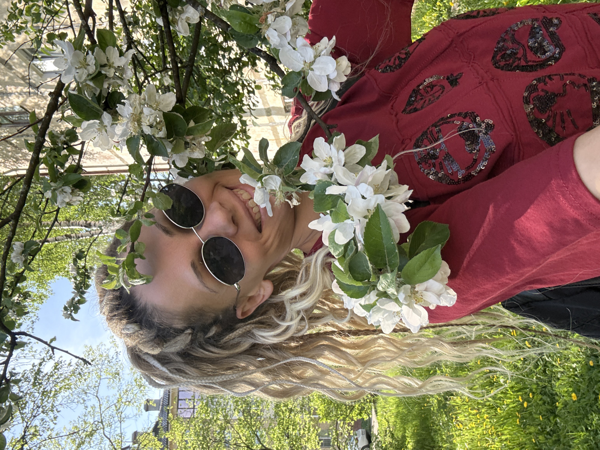A fair-skinned person with long blonde braided hair and wearing round sunglasses is smiling amongst branches of blooming white flowers. The person is wearing a maroon, sequined sweatshirt. In the background is a green lawn and some residential buildings with light-colored facades, indicating an urban area. Given the query, the location is likely Rusanovo, Russia.