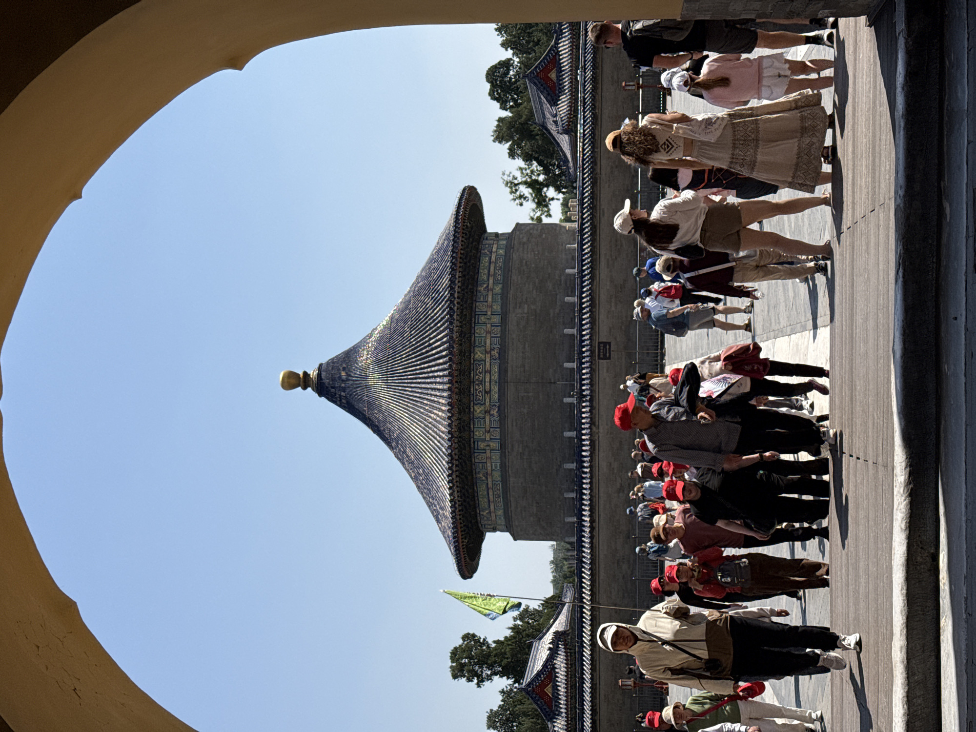 The photograph depicts a view of the Temple of Heaven in Dongcheng District, Beijing, China, as seen through an arched opening. The iconic temple structure is centrally positioned, showcasing its tiered, blue-tiled roof topped with a golden finial, and its surrounding brick wall. A large number of tourists, some wearing red hats, are seen walking on a paved path alongside the temple complex. The sky above is clear and blue.