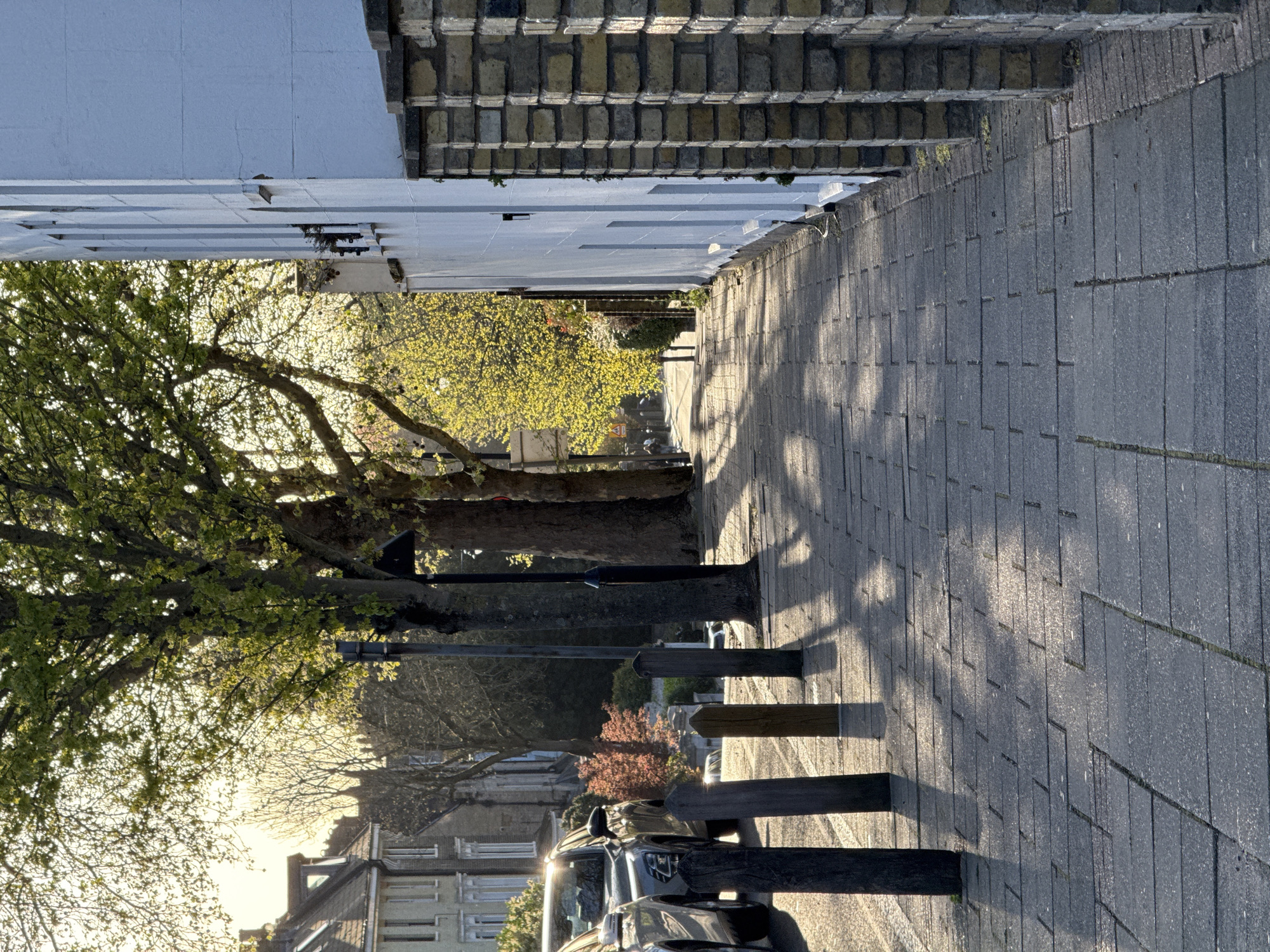 The image depicts a sidewalk in a residential area, possibly in London, United Kingdom. The sidewalk is constructed of grey paving stones. Along the sidewalk are wooden posts that appear to be bollards or a form of low fencing. Cars are parked along the side of the road, with sunlight glinting off their surfaces. Trees with green foliage line the street, casting shadows on the sidewalk. Buildings are visible on both sides of the street. The buildings range in style, with some having brick facades and others with white exteriors. The scene is illuminated by sunlight.