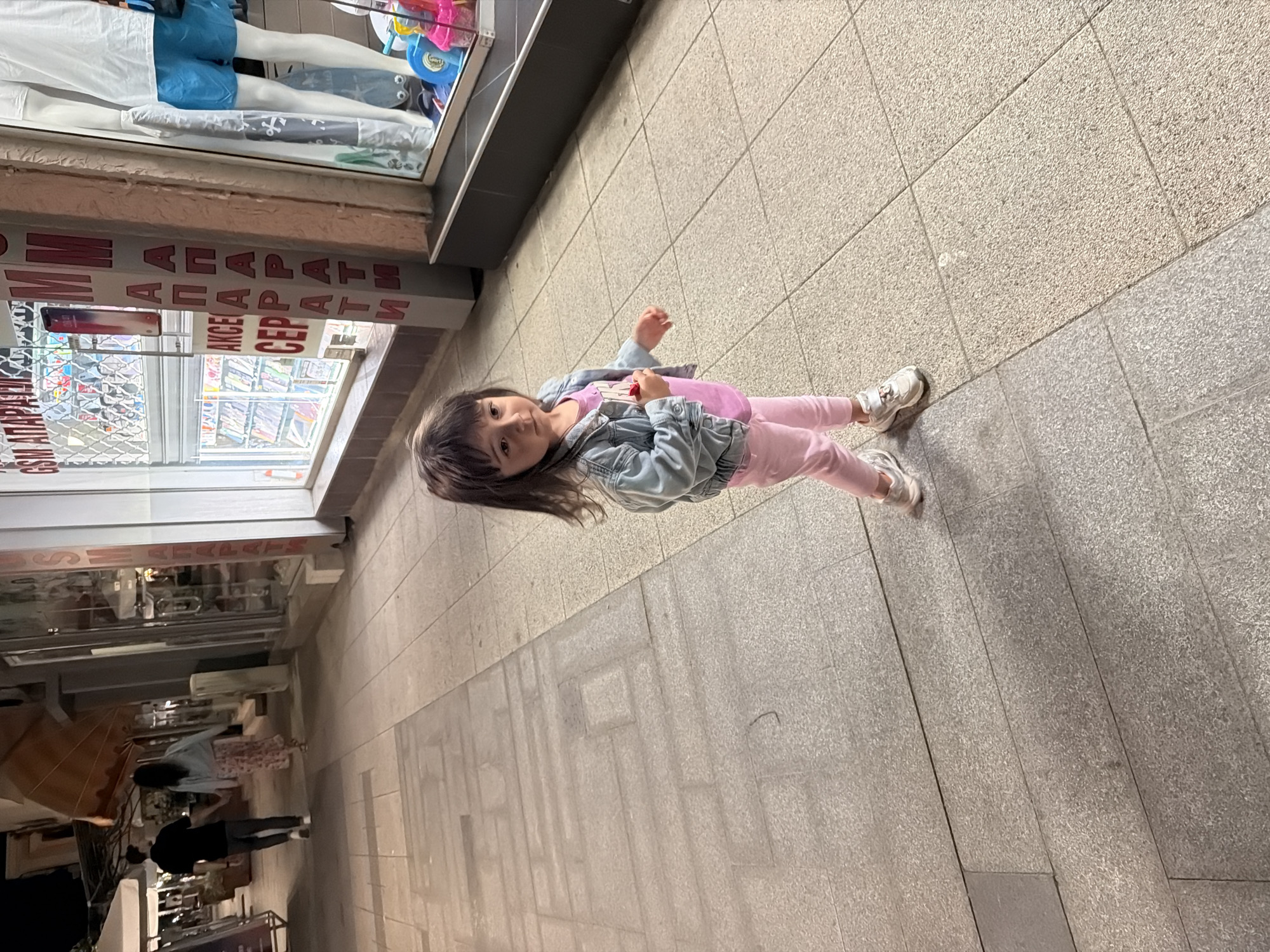 An outdoor, eye-level, medium shot shows a young girl with dark hair and bangs standing on a tiled sidewalk. She is wearing a light blue denim jacket over a pink t-shirt and pink leggings, with white sneakers. The girl is holding a small red object in her right hand and looking towards the viewer with a serious expression. Her left arm is bent at the elbow, and her hand is held in front of her.
The sidewalk is made of large, rectangular gray tiles, some of which are slightly discolored or shadowed. In the background, to the left, two figures are walking away from the camera. The person in front is wearing a black shirt and dark pants, and the person behind them is wearing a light blue jacket and a floral-patterned skirt. They are holding hands.
To the right of the sidewalk, there is a shop window with mannequins and merchandise. Vertical text on the shop facade reads "GSM АПАРАТИ" (GSM APPARATUS) and "АКСЕСОАРИ СЕРВИЗ" (ACCESSORIES SERVICE). The shop has large glass windows displaying various items, including electronics and clothing.
The overall scene appears to be on a street in Burgas, Bulgaria, likely during the evening or at dusk, given the lighting. The ground is well-lit, but shadows are present, suggesting artificial lighting. The architecture and shop signage are consistent with a commercial street in an urban environment.
