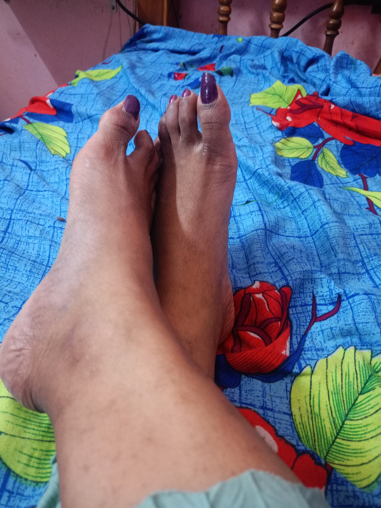 A close-up, overhead shot shows a pair of feet resting on a blue patterned bedsheet. The bedsheet has a design of red roses and green leaves. The feet are bare, with toenails painted a dark purple color. The left foot is placed on top of the right foot, with the toes of the left foot pointing towards the viewer. The background shows a pink wall and wooden furniture, suggesting an indoor setting in Chenkavila, India.