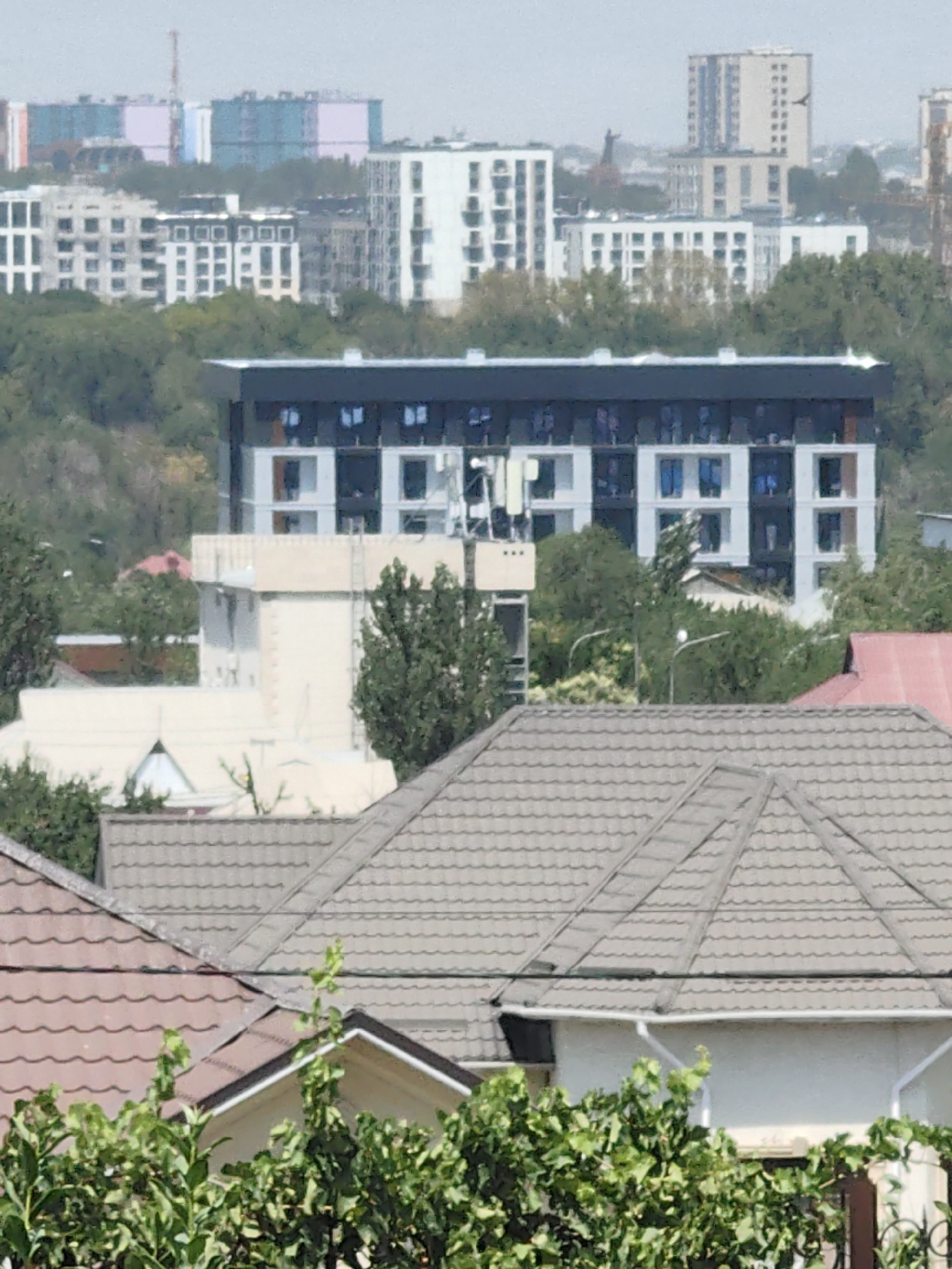 The image shows a cityscape with multiple buildings of varying architectural styles and heights. In the foreground, there are tiled roofs of residential buildings and dense green foliage. Behind these, a modern, multi-story building with a dark roof and white facade with dark-trimmed windows is prominently featured. Further in the background, several other buildings, some appearing to be under construction or recently completed, are visible, including a tall, light-colored tower. The distant skyline features larger, more colorful structures, suggesting a developing urban area. The scene appears to be set in Shymkent, Kazakhstan, based on the context provided. The environment is dominated by urban structures interspersed with green spaces, indicating a mix of residential and possibly commercial development. There are no people or animals visible in the image, and no specific actions or interactions are depicted. The overall impression is one of a contemporary urban landscape.