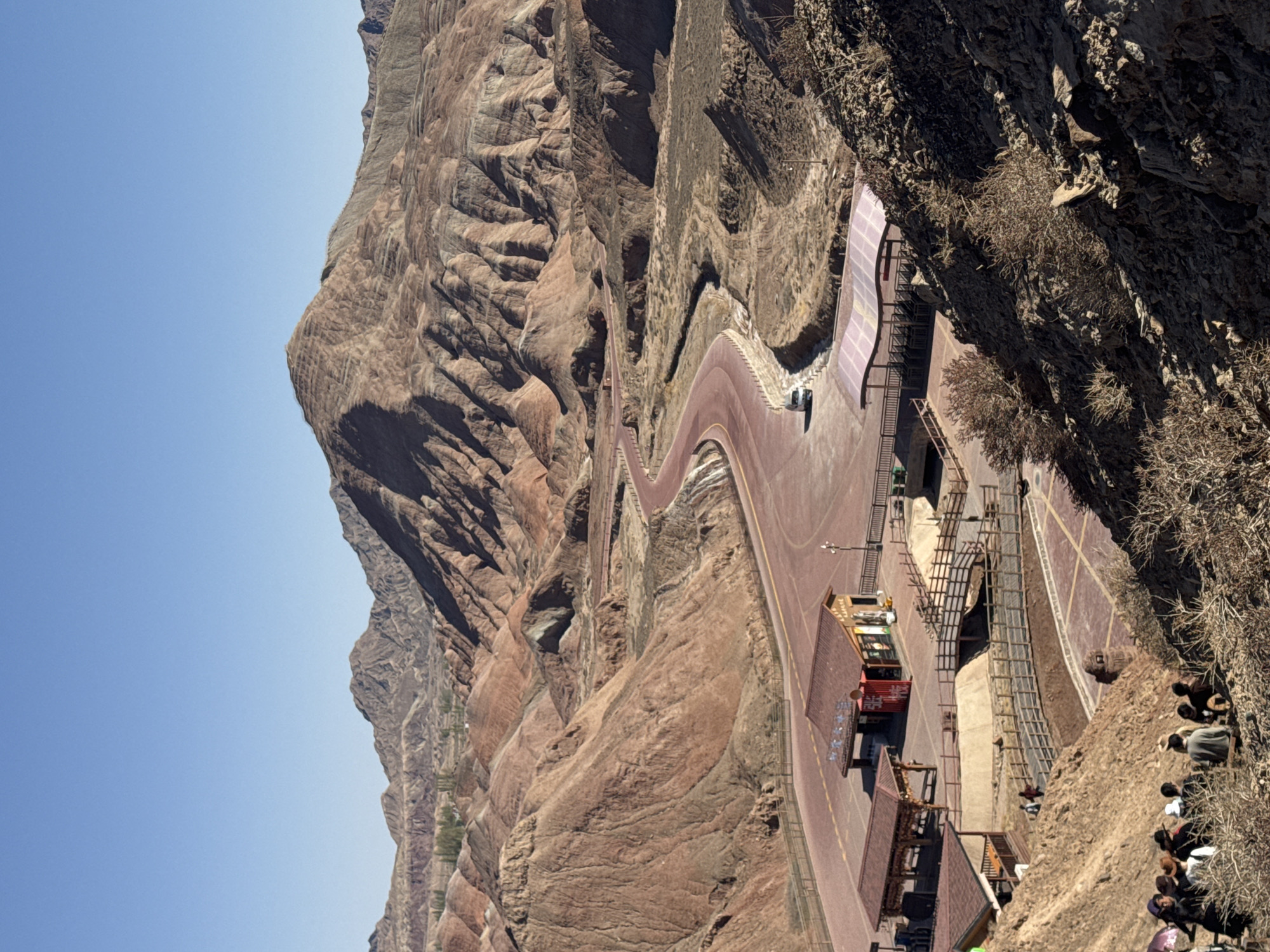 This image presents an expansive view of an arid mountainous landscape, likely a geological park in Nantai, China, under a clear, bright blue sky. The scene is dominated by massive rock formations with varied textures and colors ranging from reddish-browns and tans to grays, indicative of natural erosion and layering.
A prominent feature is a wide, reddish-brown paved road or pathway that snakes through the mountainous terrain, ascending and curving along the slopes. Adjacent to this road, a lighter-colored path or walkway runs alongside it in certain sections. Infrastructure for visitors is evident, including several small buildings or stalls with traditional-style roofs and a distinctive red container-like structure, possibly a shop or information point. Metal and wooden steps and walkways are also visible, facilitating movement across different levels of the area.
On the road, a red tourist bus or shuttle is parked, along with what appears to be a smaller yellow vehicle. To the right of the frame, a group of people is gathered on a dusty, sloping bank, some appearing to be seated or resting, observing the surroundings. Sparse, dry vegetation clings to the rocky cliffs and ground.
The image suggests a sunny mid-day with ample natural light. The overall impression is of a developed tourist attraction designed for exploring the dramatic natural beauty of the mountains. White Chinese characters are visible on the side of the red container-like building, but their specific content is not legible.