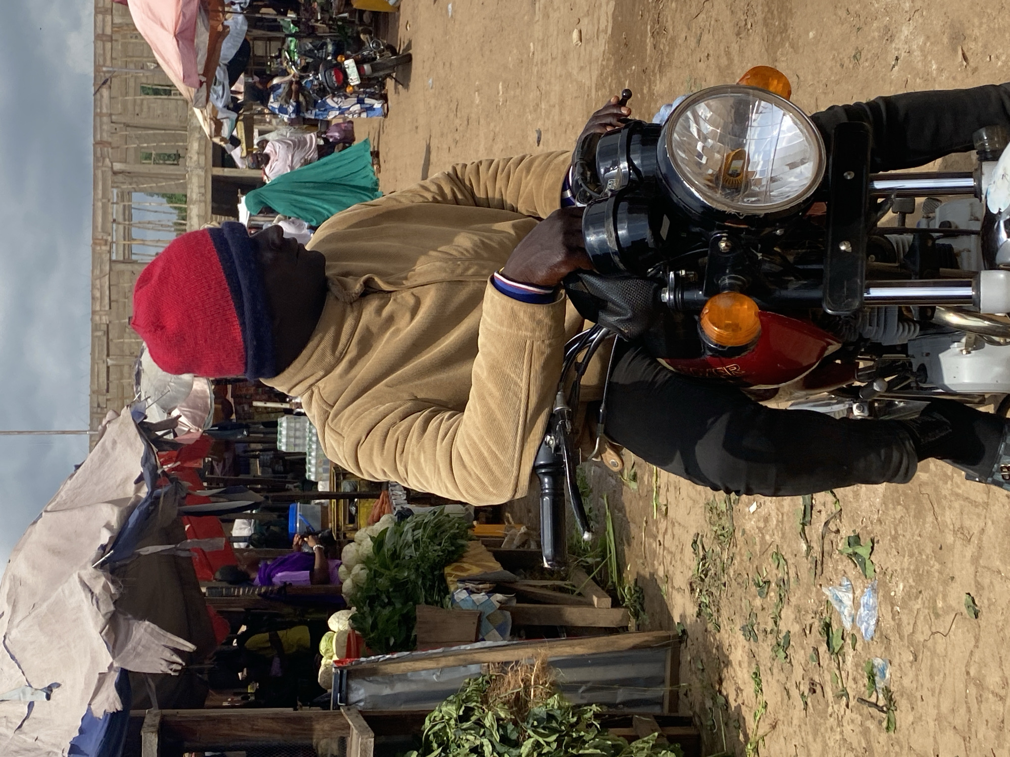 The image captures a man wearing a red and blue beanie, a tan jacket, and black pants, seated on a motorcycle. He is positioned in what appears to be an outdoor market, with stalls and various items visible in the background. The market setting includes an unfinished building under construction, tents, and produce such as cauliflower and leafy greens displayed on wooden crates.
The man is looking to his right, with his hands on the handlebars of the motorcycle. The motorcycle itself is a prominent object, with its headlight and orange turn signals clearly visible. The ground is dirt and scattered with some debris.
The scene suggests a daytime setting, possibly mid-morning or afternoon, given the illumination and shadows. The weather appears to be clear. The overall atmosphere of the image is that of a bustling marketplace, with the man and his motorcycle being a central focus. The presence of the construction site might indicate ongoing development or renovation in the area. There is no visible text in the image. The man's expression is neutral. The location context is Kafanchan B, Nigeria.