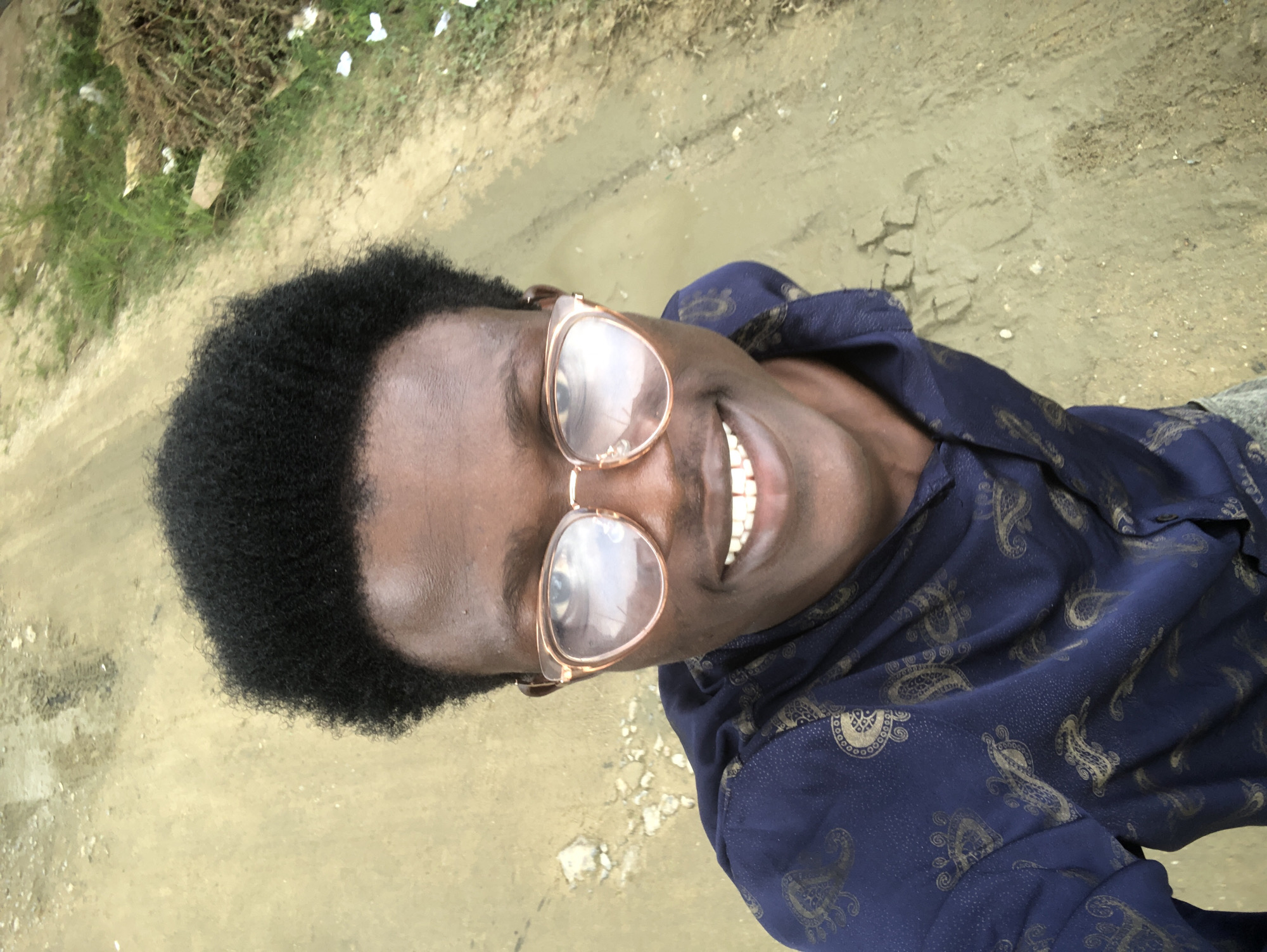 A young Black male is taking a selfie outdoors. He has dark skin, short black afro-style hair, and is wearing large, round, tan-rimmed glasses. He has a wide smile, showing his white teeth. He is wearing a dark blue shirt with a gold paisley pattern. The background is a dirt road, with some green grass and dry brush to the right. The scene appears to be in a rural or semi-rural area, possibly on the outskirts of Ado, Nigeria, based on typical landscapes and residential areas found there.