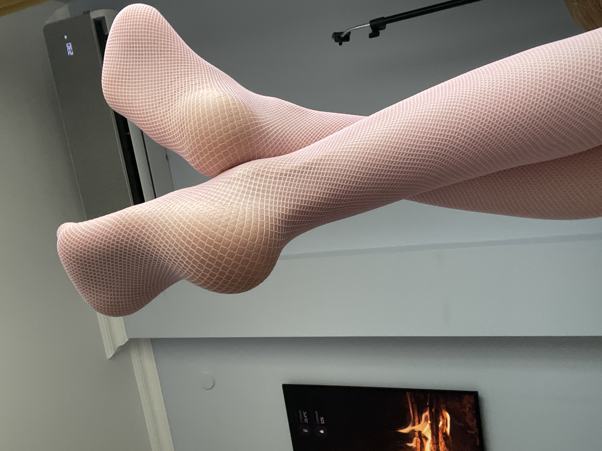The image shows a close-up view of a person's legs and feet, wearing pink fishnet stockings. The legs are crossed and positioned in a way that highlights the texture of the stockings. In the background, an air conditioning unit with a digital display reading "23.0" is visible on a light blue wall. To the left, a framed digital display shows an image of a fireplace with flames and displays text including "23.4°C" and "52%". A thin black pole, likely part of a tripod or stand, is visible on the right side of the image. The overall setting appears to be an indoor environment, possibly a bedroom or living area. The image does not provide any specific information that would allow for identification of the location within the city of Yablonovskiy, Russia.