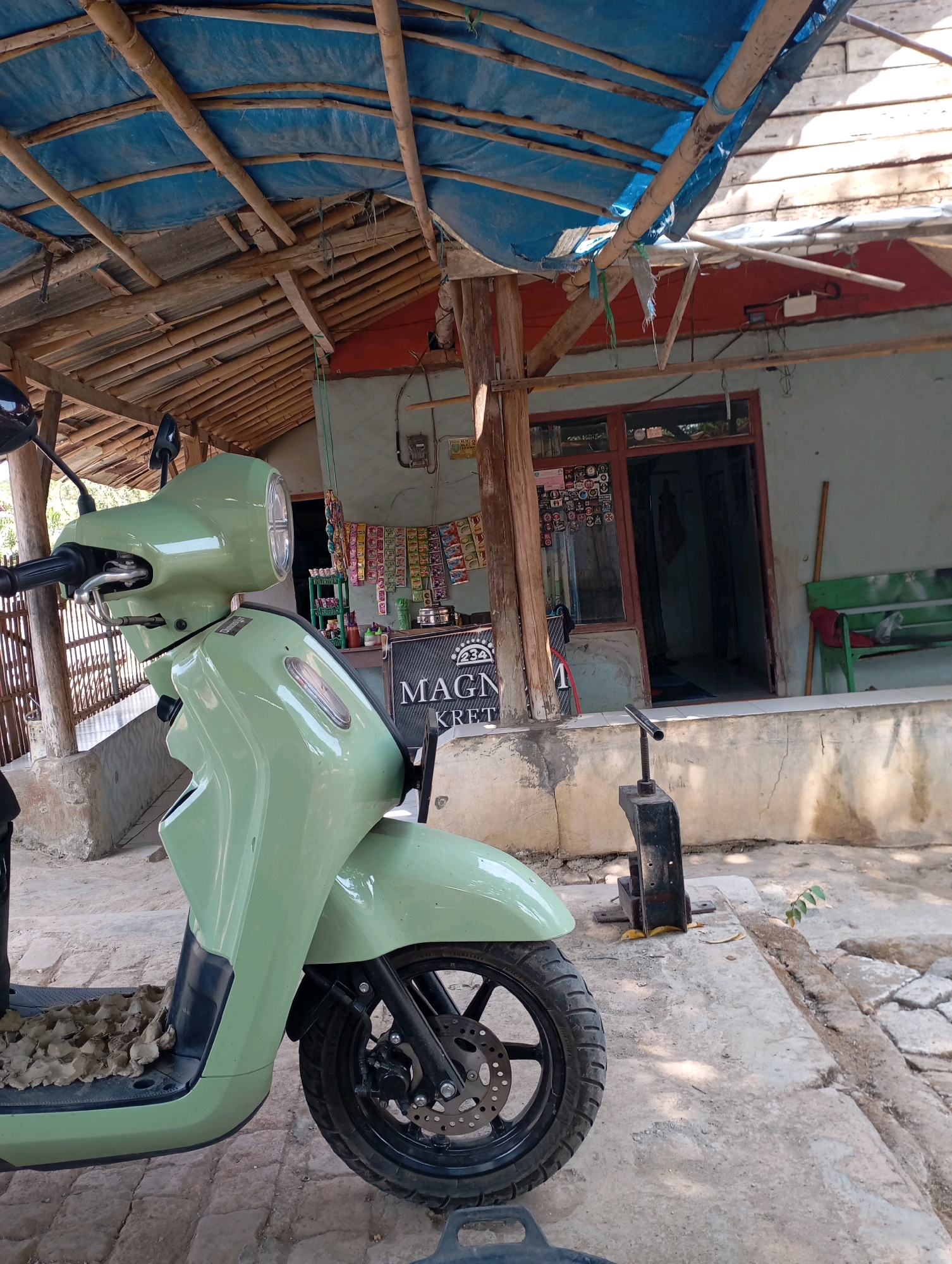 The image captures an outdoor scene in Kecamatan Haurgeulis, Indonesia, focusing on a light green scooter parked in front of a building. The building appears to be a simple structure with a red wall and a wooden door with glass panes. Above the building, a blue tarp is stretched over a bamboo frame, suggesting a temporary or makeshift roof. The ground is paved with concrete and some cobblestones.
The scooter is the primary subject, positioned in the foreground and taking up a significant portion of the frame. Its front wheel, fender, handlebars, and part of the body are visible. The footrest area of the scooter is covered with what looks like cardboard egg cartons. Behind the scooter, a black metal object, possibly a tool or a stand, is visible on the concrete.
To the left of the scooter, a wooden fence or partition is present. To the right of the scooter and behind the building, a green wooden bench is situated. The building's facade displays various items, including what appear to be packaged snacks or goods arranged on shelves and a sign with text that is partially obscured but includes "MAGNA KRET". Electrical wiring and a meter box are also visible on the exterior wall of the building. The overall environment suggests a residential or small commercial area. No people are visible in the image.