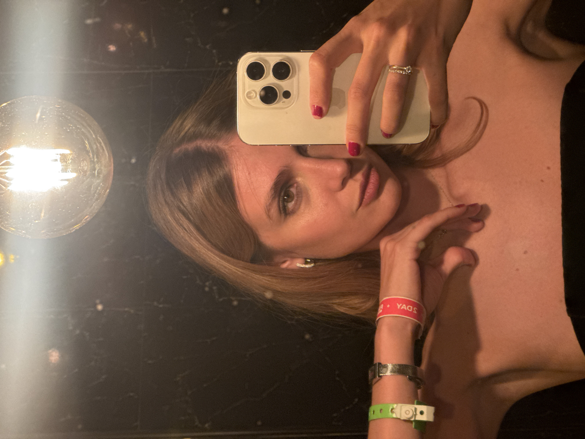 A medium shot depicts a woman in Yerevan, Armenia taking a selfie in front of a mirror, with an incandescent lightbulb visible in the upper left. She holds a white smartphone, covering the top part of her forehead, with a hand sporting a diamond ring on the ring finger and magenta nail polish. The woman has light skin, light brown hair, and is wearing what appears to be a black top or dress. Her left wrist has a red wristband with the word "YAGS," a silver watch, and a green and white wristband. The background features a dark, reflective surface, presumably the mirror she's using to take the photograph.