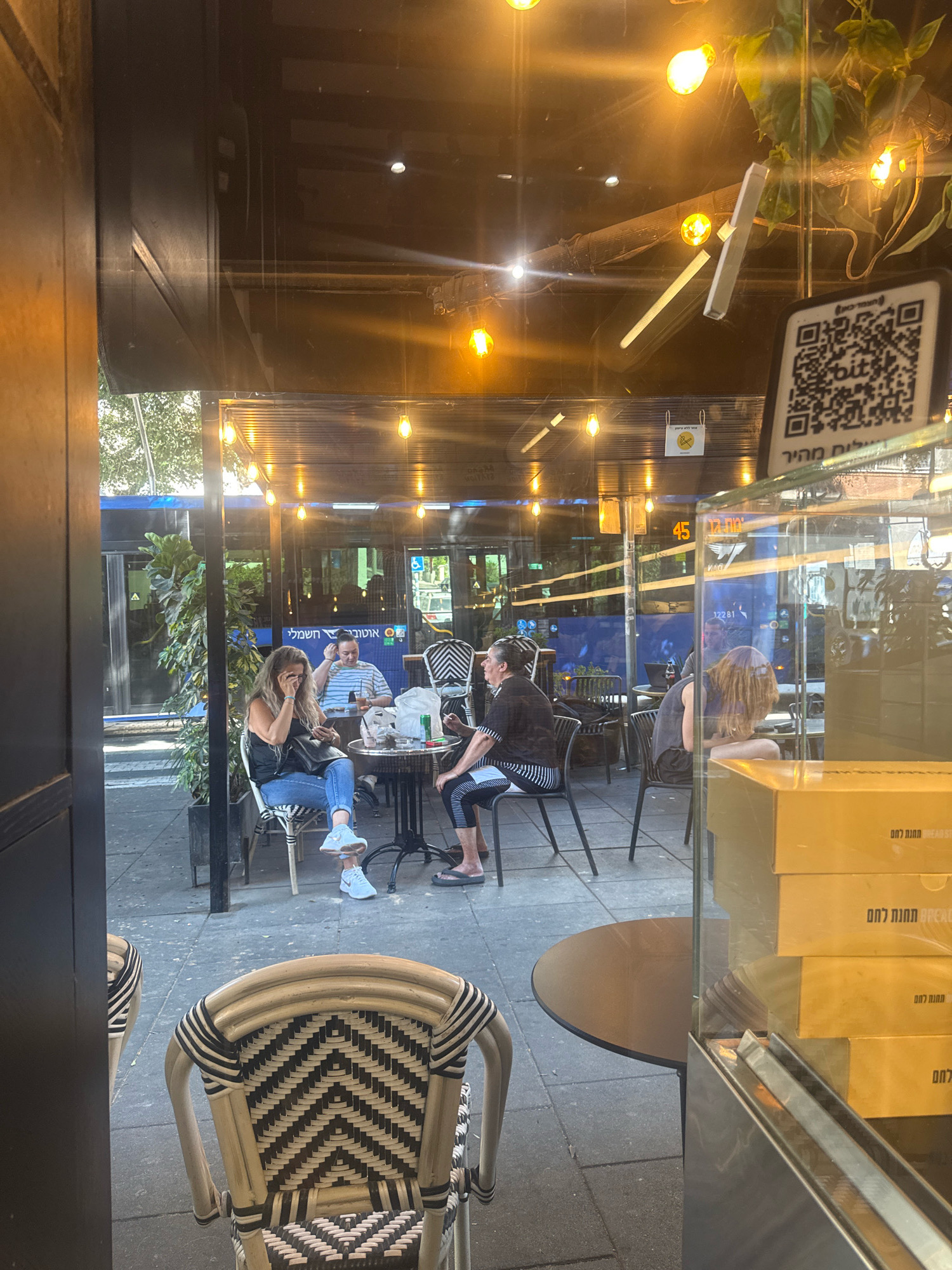 This is a street-level, eye-level, medium shot photograph taken from the interior of a cafe or restaurant, looking outwards through a glass partition and windows. The scene depicts an outdoor seating area of a cafe on a sunny day.
In the foreground, a wicker chair with a black and white chevron pattern is visible on the left. To the right, a glass display case contains stacked cardboard boxes. Text in Hebrew is visible on the boxes, with one box also showing English text "BREADST".
The outdoor seating area is furnished with tables and chairs. Three people are seated at a table. A woman with blonde hair in a black sleeveless top and blue jeans is seated on the left, looking at her phone. A man in a striped t-shirt is seated in the center, looking towards the woman. Another person, with red hair and wearing a dark sleeveless top, is seated on the right, facing away from the camera and leaning forward. There is food and drinks on the table.
Behind the seating area, a blue bus is visible, suggesting the cafe is located near a street or transportation hub. The environment appears to be a city, with trees and foliage visible in the background. The lighting suggests it is daytime, with sunlight filtering through the cafe's roof and illuminating the scene. A QR code is displayed on the glass partition, likely for payment or information.
The location is identified as Ramat Gan, Israel.