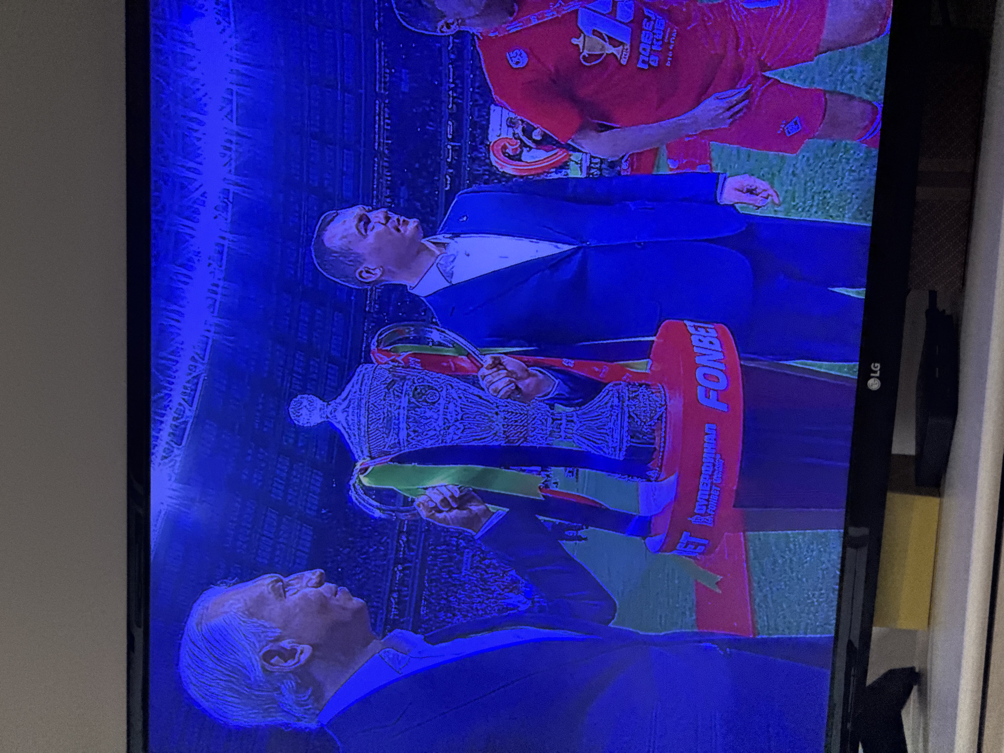 The image shows a television screen displaying a scene from what appears to be a soccer event, likely in Izhevsk, Russia. On screen, three men are visible. Two of the men are holding a large trophy with the word "FONBET" prominently displayed on it. The men are wearing suits, and a third man in a red jersey with "POBEA B KY6" visible, stands slightly behind them. The backdrop shows a stadium filled with spectators. The surrounding environment shows the edge of the television and the surrounding wall.