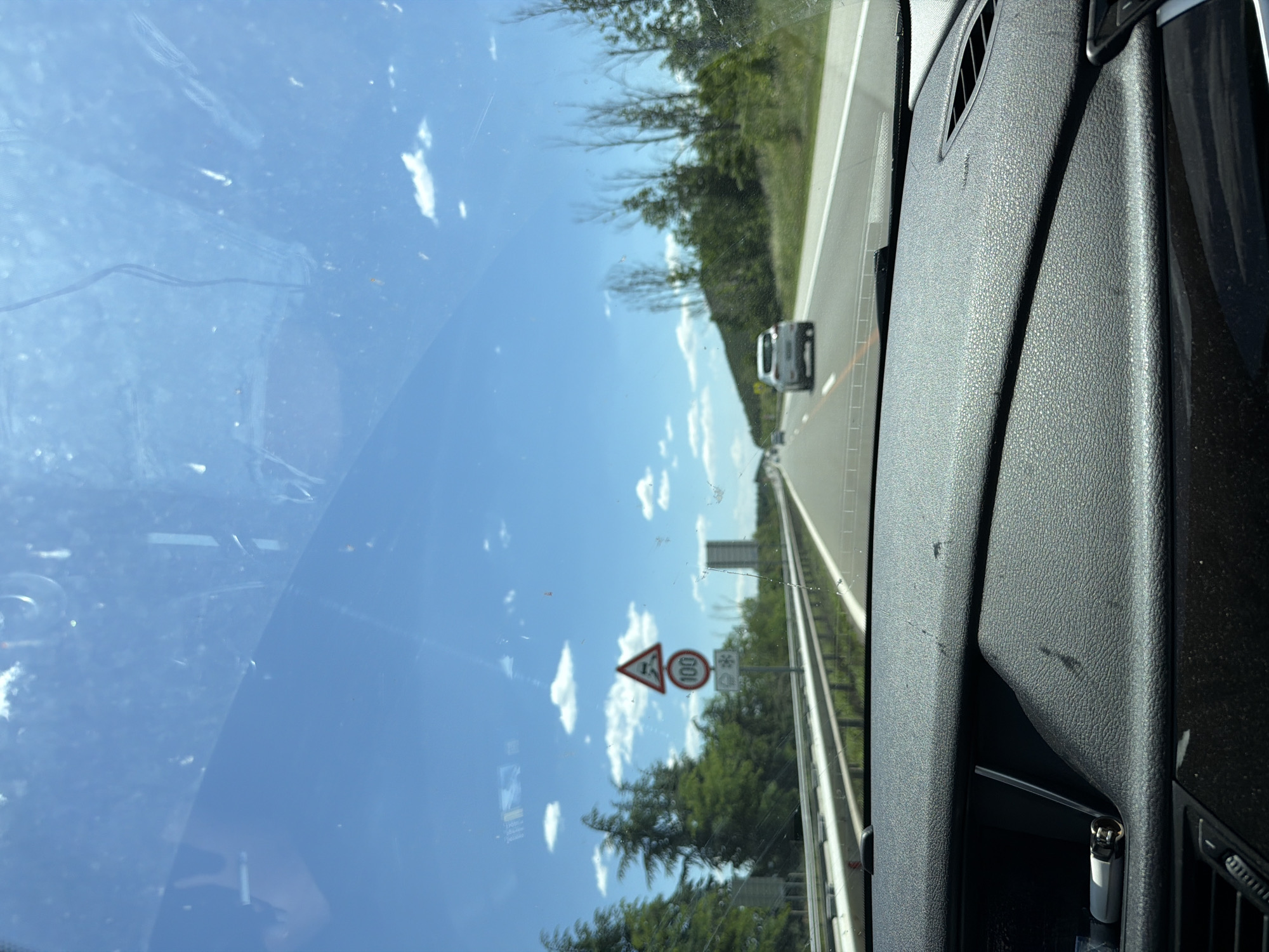 A view from inside a car driving on a highway. The driver is traveling on the right lane. A white car is visible ahead in the same lane. A speed limit sign indicating 100 is visible on the right side of the road, along with a hazard warning sign and a snow warning sign. The sky is blue with scattered clouds. The location is along a highway, and there is no evidence that this is Slavovitsa, Bulgaria. The interior of the car, including the dashboard and vent, is visible on the right. A lighter is visible in a compartment within the dashboard. The windshield has some smudges.