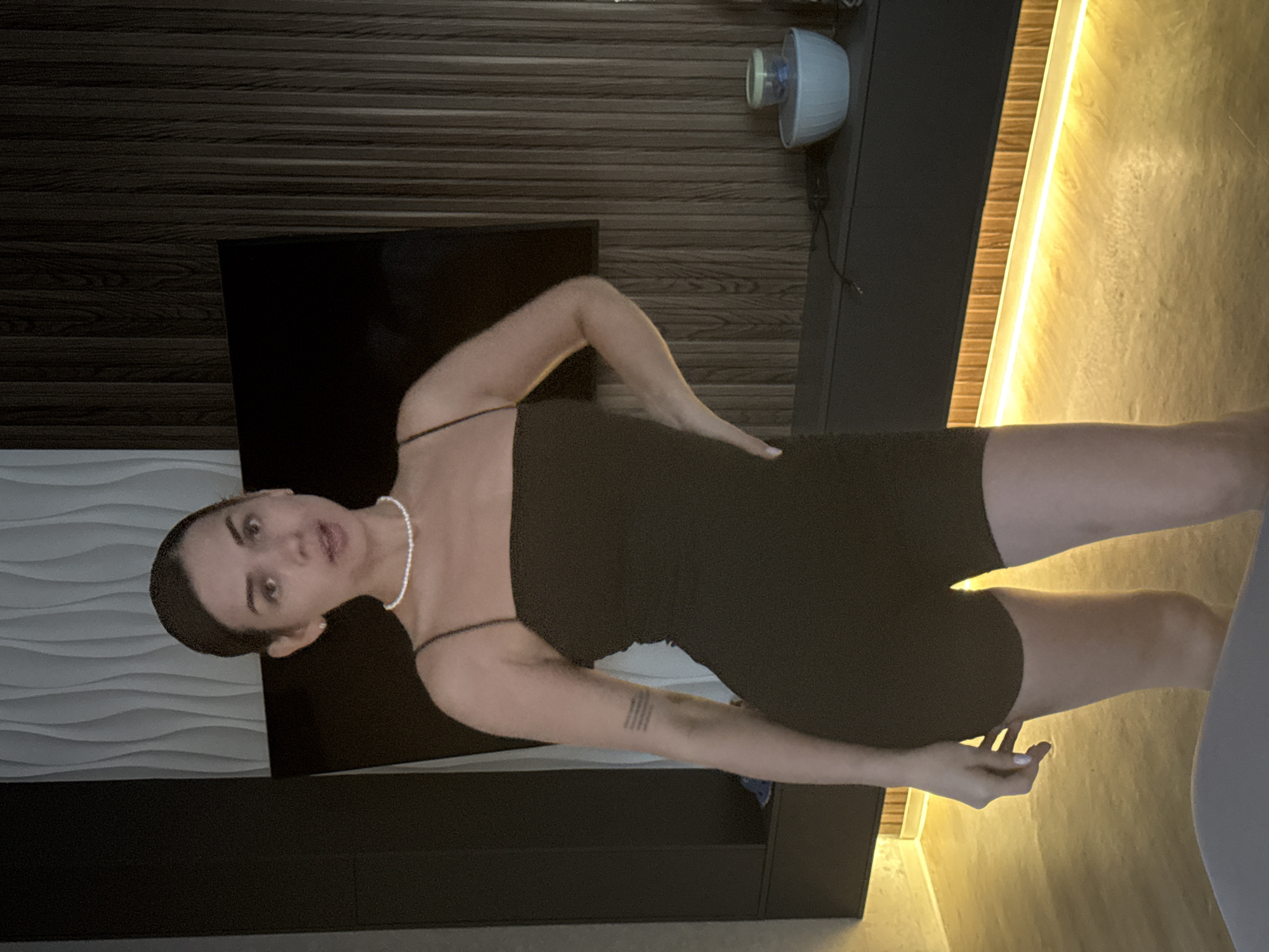 A woman is standing in a room. She is wearing a black sleeveless dress and a pearl necklace. Her right hand is on her hip, and her left hand is by her side. The background includes a textured wall, a television, a cabinet, and a light fixture. Based on the query, the location is Лобаново, Russia.