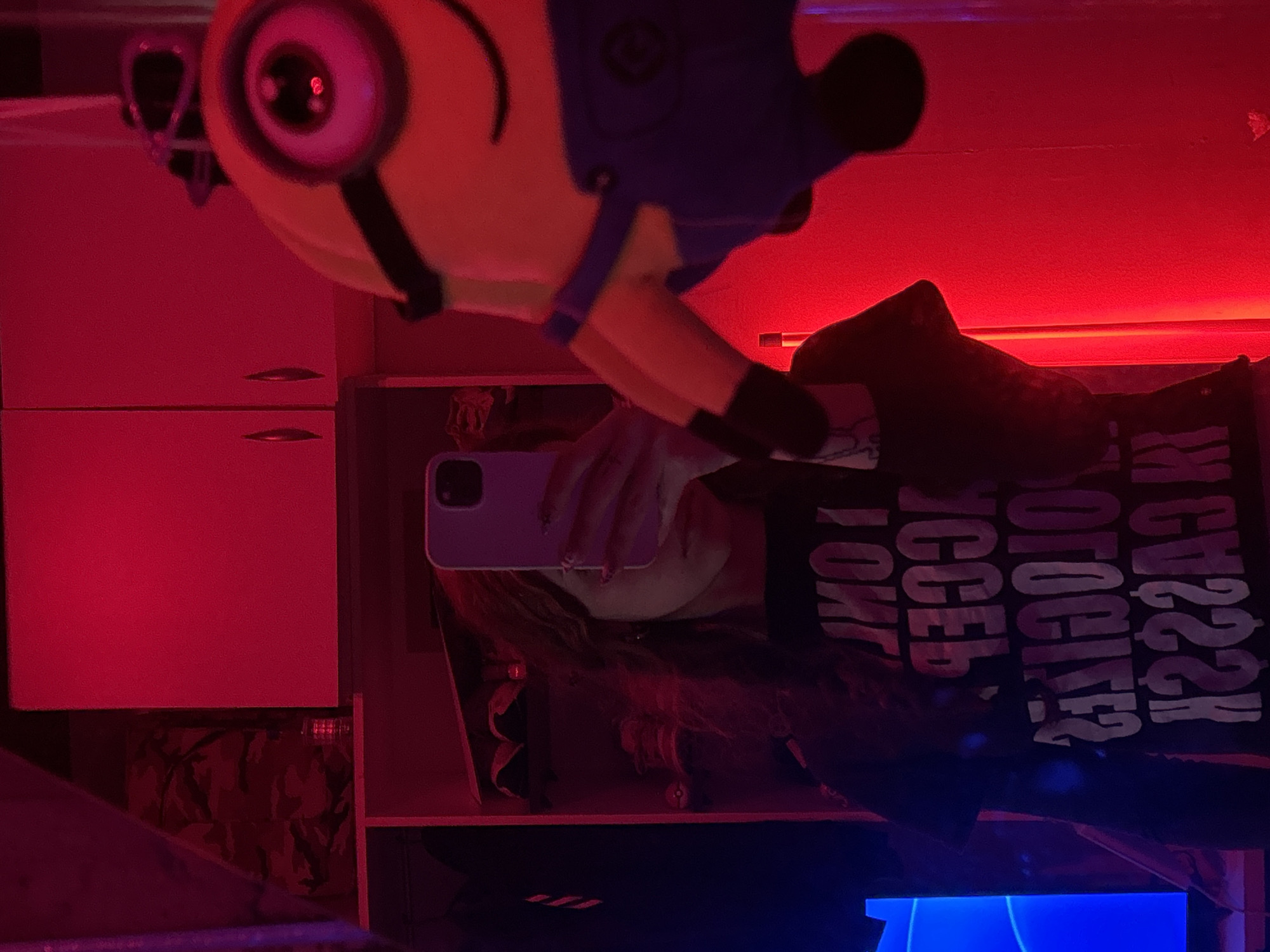 A selfie-style image shows a person holding a phone in front of their face, presumably taking the picture. The person has long hair and is wearing a black shirt with white lettering that is partially obscured and upside-down. Above the person is a Minion plush toy. The background includes white cabinets and shelves. The lighting is primarily red, with some blue light at the bottom of the frame. The location is not identifiable.