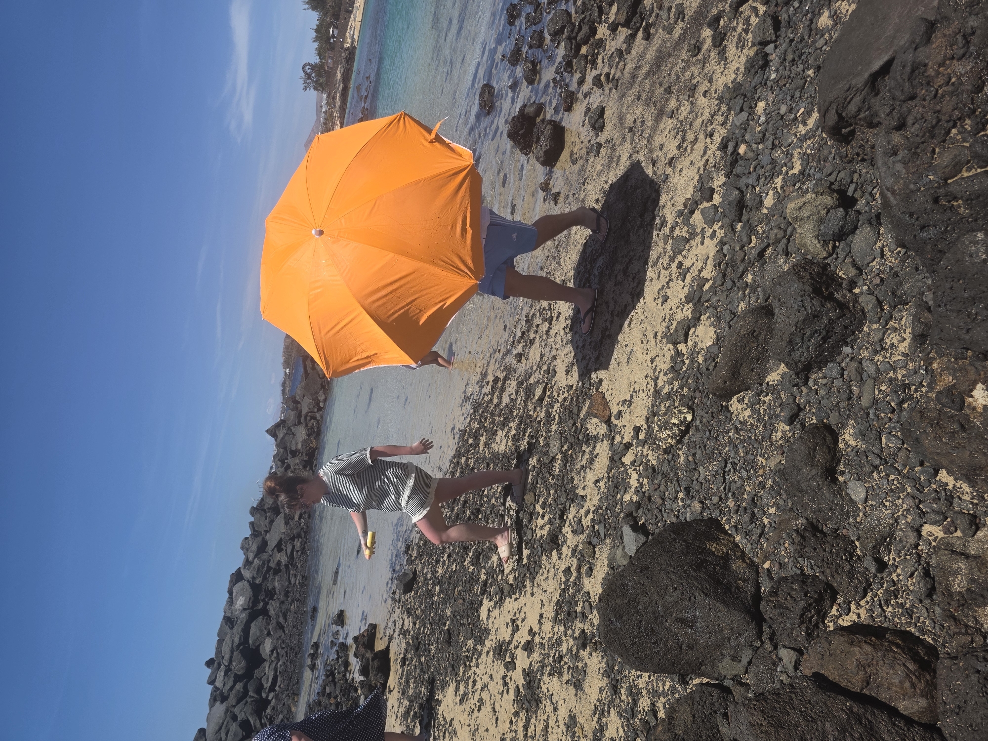 The image depicts a sunny day on a rocky beach in Costa Teguise, Spain. Two individuals are partially visible, walking near the shoreline. In the foreground, a woman with her hair in a bun is seen from the waist down, wearing a striped top and shorts, holding a yellow object. Behind her, another person is walking while holding a large, vibrant orange umbrella over their head, providing shade. The beach is composed of sand and dark volcanic rocks. The water is a clear turquoise, and the sky is a brilliant blue with a few wispy clouds. In the background, a rocky breakwater extends into the sea, and some vegetation and buildings are visible along the coast.