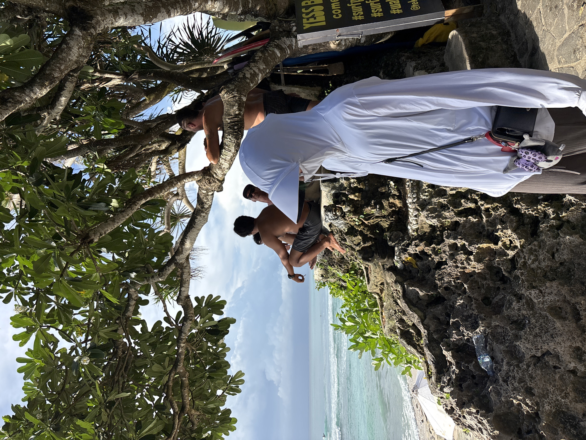 The image shows a scene at a cliffside spot in Kuta Selatan, Indonesia. A man in a white robe and sunglasses sits on a wooden bench. Next to him are a black purse and a plush toy with purple leopard spots. Further back, two men are perched in the branches of a tree overlooking the ocean. One man is shirtless and holding a small, dark object in his hand. The ocean is visible beyond the cliff, with a light blue color under a partly cloudy sky. The area is surrounded by trees and tropical vegetation growing from the rocky cliffside. A sign with text that includes "#surf" is visible above the men in the tree. A plastic water bottle can be seen embedded in the rocks.