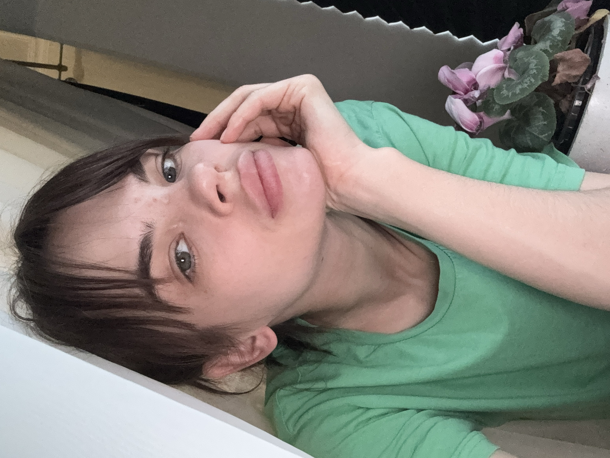 A woman with brown hair and bangs, wearing a green shirt, is lying down with her head resting on her hand. She is looking up at the camera. A potted plant with pink flowers is visible in the upper right corner of the frame. The background includes a window and a white surface. The image's location in Rostov-on-Don, Russia, is not determinable from the provided visual information.