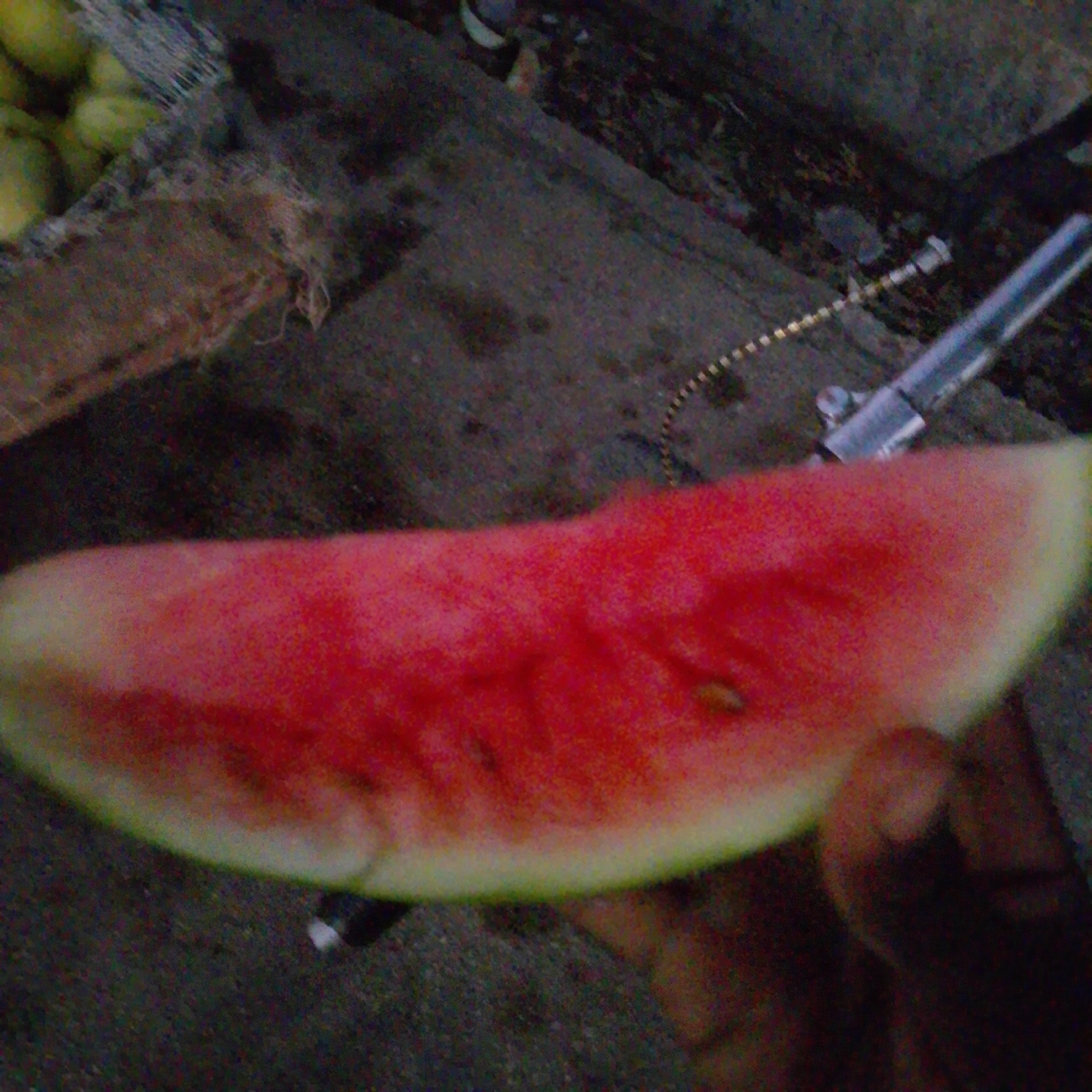 The image shows a close-up of a slice of watermelon. The watermelon is bright red with some visible seeds and a pale green rind. A person's hand is partially visible on the right side of the image, holding the watermelon. In the background, there is a rough, dark surface, possibly concrete or asphalt, with some debris scattered on it. To the left of the watermelon, there appears to be a brown, textured material, possibly burlap or a woven bag, and some round, yellow objects that could be other fruits or vegetables. A metal object, possibly a part of a tool or equipment, is also visible in the upper right portion of the background, with a thin, coiled wire extending from it. The overall lighting suggests it could be daytime, but the quality of the image makes it difficult to determine precise details about the environment or the time of day. The setting appears to be an outdoor, possibly a market or street stall, given the presence of produce and the rough ground. There is no visible text in the image.