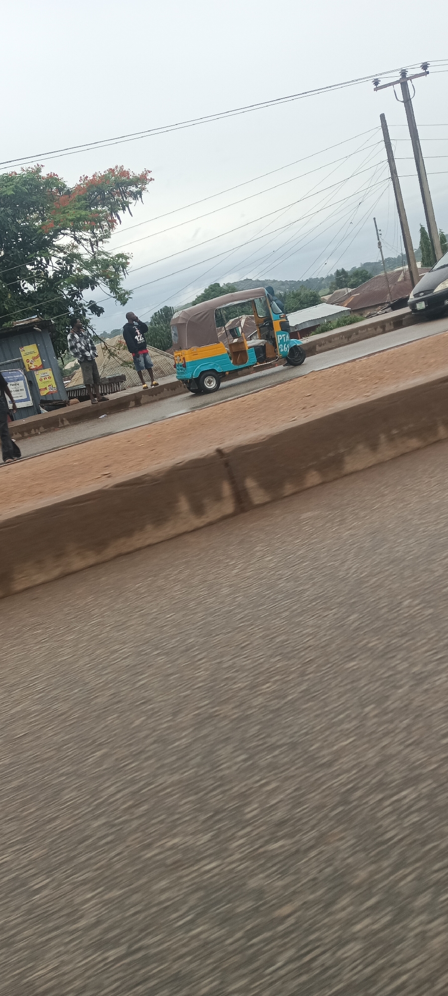 The image presents an overcast street scene in Bukuru, Nigeria, captured from a slightly elevated perspective, likely from a moving vehicle, as indicated by the motion blur on the road surface in the foreground.
In the midground, a colorful three-wheeled vehicle, commonly known as a keke napep or tuk-tuk, is prominently featured. It has a light blue and yellow body with a brown canvas canopy, and a driver is visible inside. On its side, the text "PTA 269" is clearly visible in blue lettering.
To the left of the tricycle, two men are standing on a raised concrete sidewalk. One man, dressed in a plaid shirt and shorts, faces towards the tricycle. Another man, wearing a dark hoodie and shorts, stands nearby. Further left, a third person is partially visible walking past a small roadside stall with corrugated metal walls. This stall displays several posters, one of which faintly shows text that appears to be "DATA" or "DATA BUNDLES."
The background features multiple utility poles supporting numerous overhead power lines stretching across the sky. Green trees, including one with distinctive red-orange flowers, are visible, along with distant hills and scattered buildings with dark roofs, all under a grey, cloudy sky. A black car is partially visible on the far right of the road. A low concrete barrier separates the road from the pedestrian area where the people are standing. The scene portrays a typical urban or semi-urban setting with daily activities and local transportation.