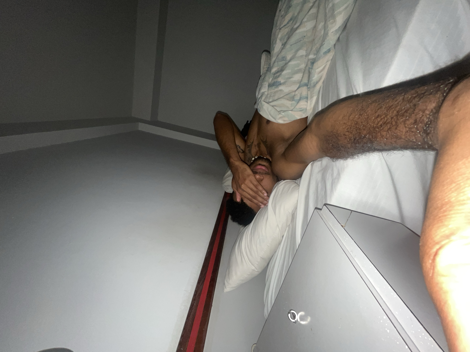 The image shows a person lying in a bed, with their face covered by their hands. The person is wearing patterned pajama bottoms. The scene is dimly lit, suggesting it is nighttime or the room is dark. The setting appears to be a bedroom with white bedding and a white pillow. To the right of the bed, a portion of a gray surface, possibly a dresser or a cabinet, is visible. The walls of the room are light-colored. The person has tattoos visible on their chest. The overall mood of the image is one of discomfort or distress, given the way the person is covering their face.