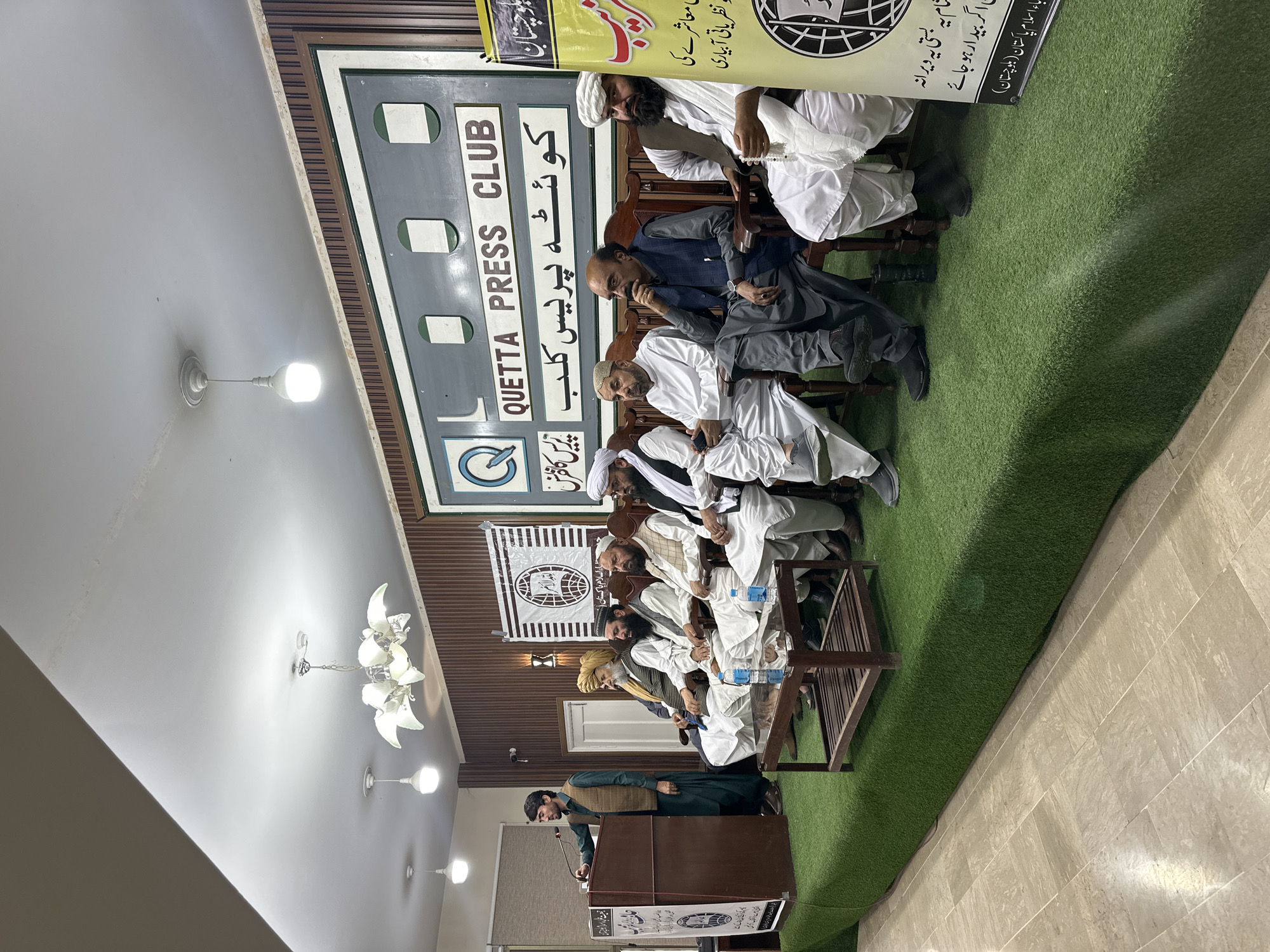 A meeting or press conference is taking place in Quetta City Tehsil, Pakistan at the Quetta Press Club. Men dressed in traditional clothing are seated on chairs. A man is at a podium speaking into a microphone. A sign in Urdu and English identifies the location as the Quetta Press Club. Additional banners in Urdu are also present. The room has a wood-paneled wall and artificial turf on the floor near the seating area.