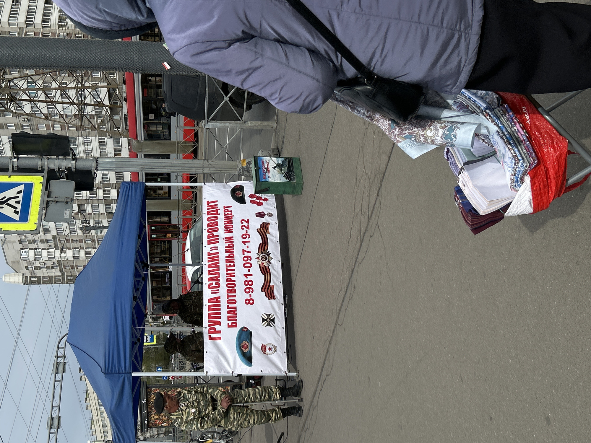 The image shows a street scene in Saint Petersburg, Russia, with a blue canopy tent set up on the sidewalk. Three men in military camouflage uniforms are standing behind the tent. A sign is hung on the tent with red text in Russian that reads "GROUP SALANG CONDUCTS CHARITABLE CONCERT 8-981-097-19-22". Below the text are images of military and Soviet symbols, including a military medal, a St. George ribbon, and a military beret. A small green box is on the sidewalk displaying a picture of soldiers and the words "We are Russian! God is with us!". A person wearing a light purple jacket stands in the foreground. To the right of the person are several stacks of folded fabric in a red bag. A traffic light and crosswalk sign are visible in the background, with apartment buildings across the street. A red and white tram also appears to be stopped in the background.