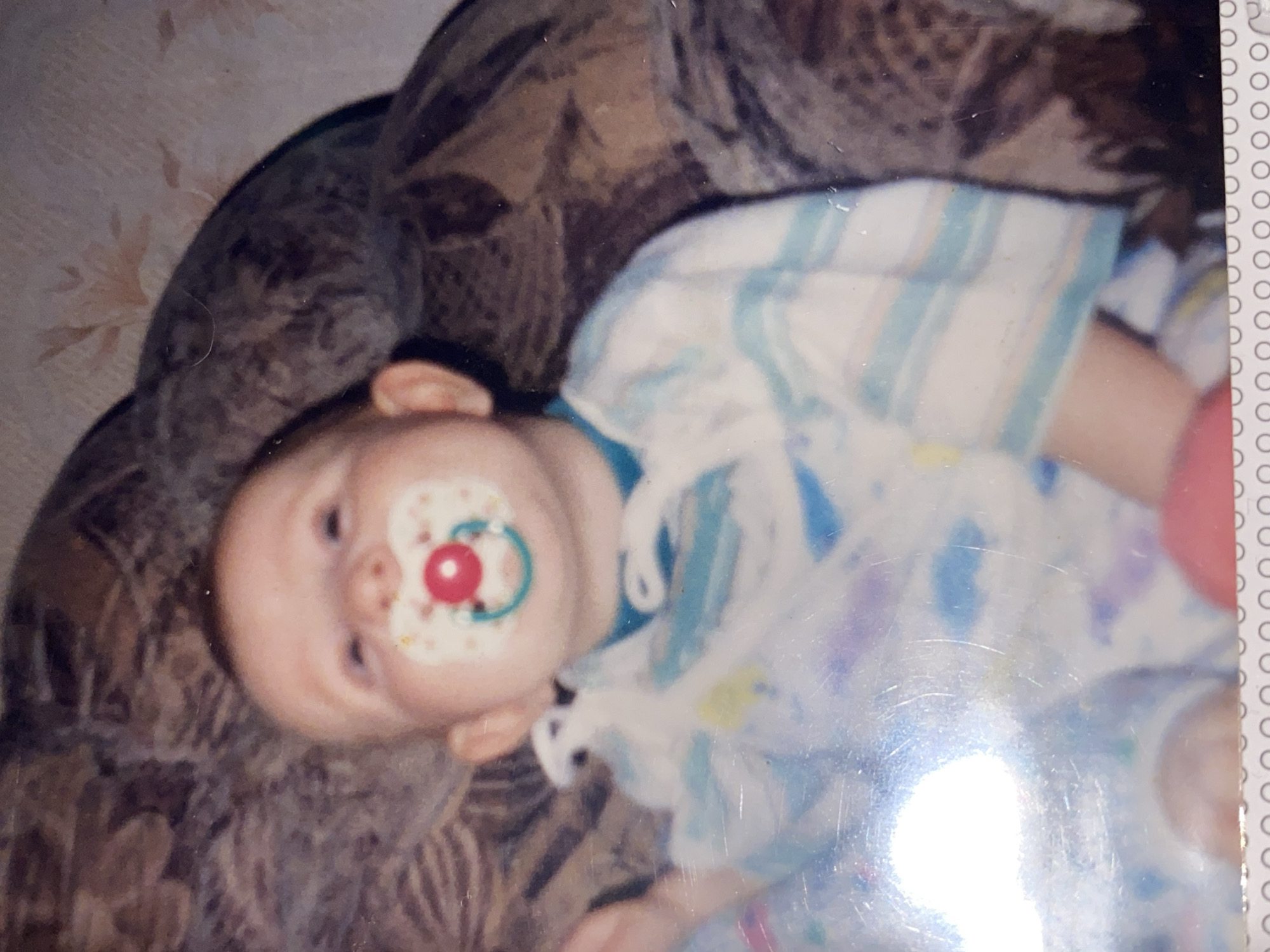 A forensic examination of the image reveals an infant subject, positioned indoors within the city of Chaikovsky, Russia. The precise address or specific room is not ascertainable from the visual data. The infant, displaying fair skin and light-colored hair, is oriented with its face directed generally upwards, engaging the camera. A pacifier is present in the infant's mouth; it is multi-colored, featuring a white shield with small yellow and purple patterns, a prominent red center button, and a teal-colored handle. The infant is attired in a light-toned, long-sleeved garment, characterized by horizontal light blue and white stripes over the upper torso, with additional faint, light-colored patterns. The immediate background consists of a dark brown and beige patterned fabric, indicative of upholstered furniture or bedding. A distinct light reflection is observed in the lower-left quadrant of the image. A portion of the infant's left arm and a segment of a pink object are visible at the bottom of the frame. No other persons or primary subjects are identifiable.