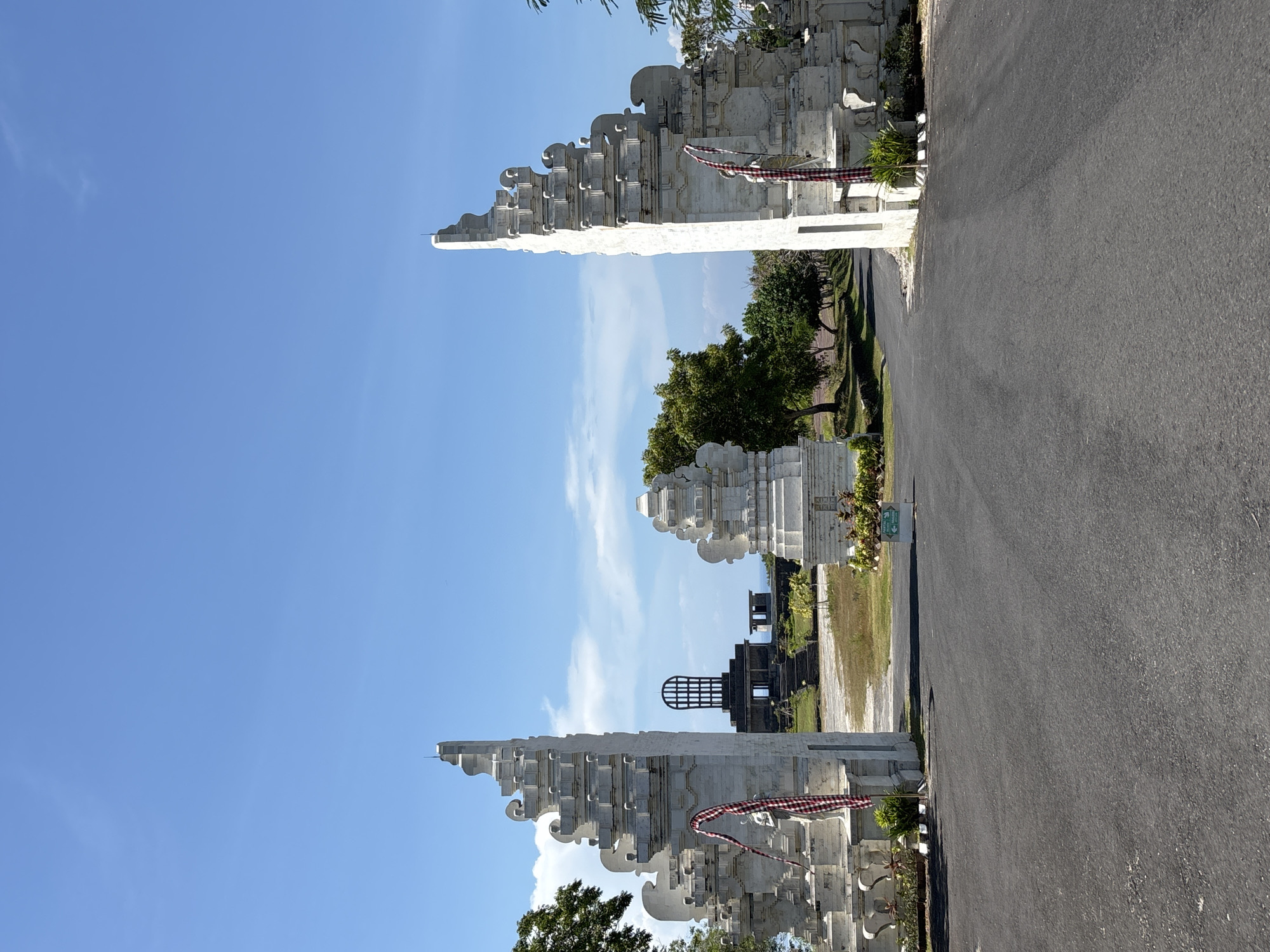 The image shows a traditional Balinese gate, known as a "Candi Bentar," located in Kuta Selatan, Indonesia. The gate consists of two symmetrical, white-stone structures with stepped, ornamental tops. Each structure has a checkered red and white cloth draped across its front. A paved road stretches between the two structures, leading into a park-like area with trees and another structure that appears to be a viewing platform or tower. The sky is blue with scattered white clouds. The lighting suggests it is daytime. There are no people visible in the image.