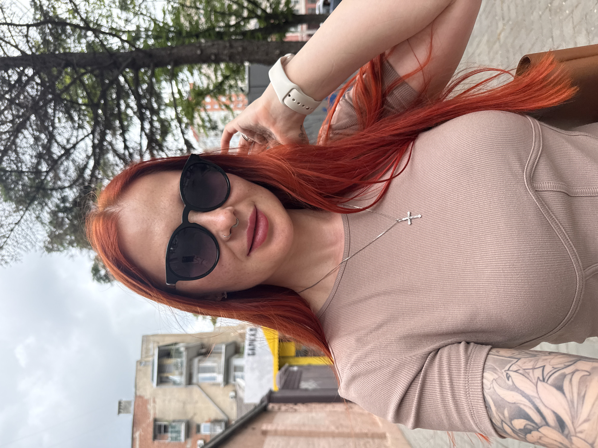 A woman with red hair is taking a selfie on a street in Rostov-on-Don, Russia. She is wearing sunglasses, a beige top, and a silver cross necklace. She has a watch on her left wrist and tattoos on her left arm. The background includes buildings, trees, and a cloudy sky.