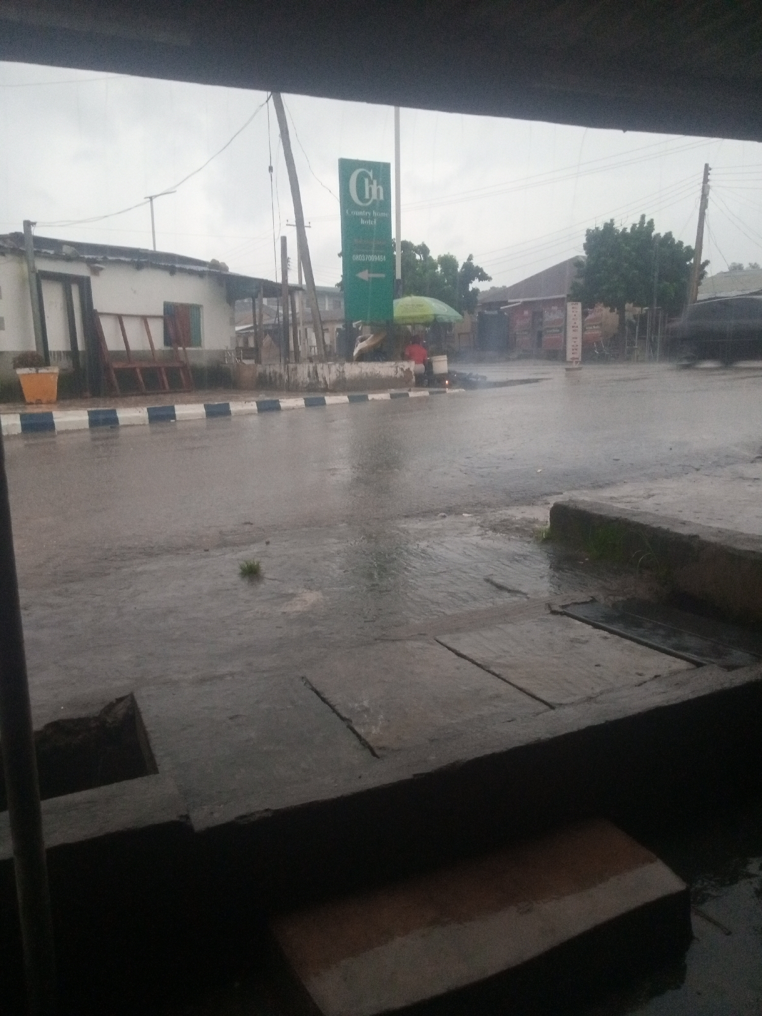 The image shows a street scene in the city of Jos, Nigeria, during what appears to be a rainy day. The road is wet and shows signs of pooling water. To the left, there is a white building with a white and blue striped curb in front of it. A tall green signpost with "Ch Country Home Hotel" written on it is visible in the background. To the right of the signpost, there are trees and other buildings. There is a person under a green umbrella in the middle of the scene, and it appears to be raining, with water droplets visible in the air. The foreground shows a dark concrete structure with a visible opening. The overall atmosphere is somber due to the overcast sky and rain.