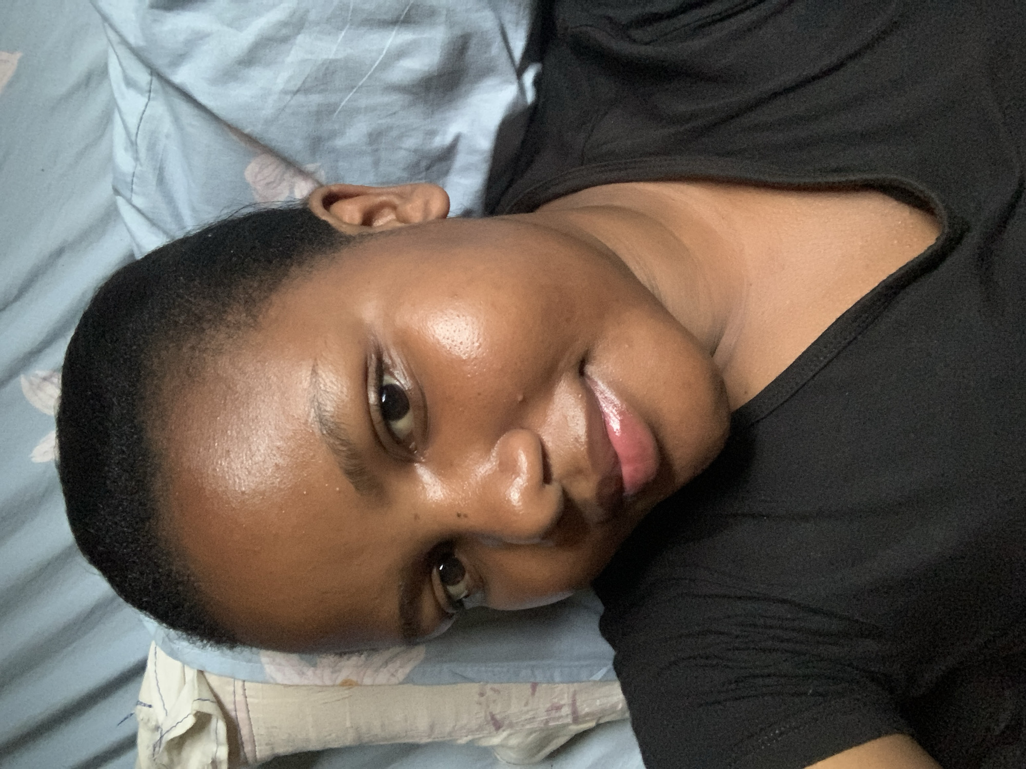 A person with dark skin, dark eyes, and short dark hair is lying on a blue patterned bedsheet wearing a black t-shirt. The person is facing the camera with a slight smile. The person is likely indoors. The location, Dar es-Salaam, Tanzania is not discernible from the image.