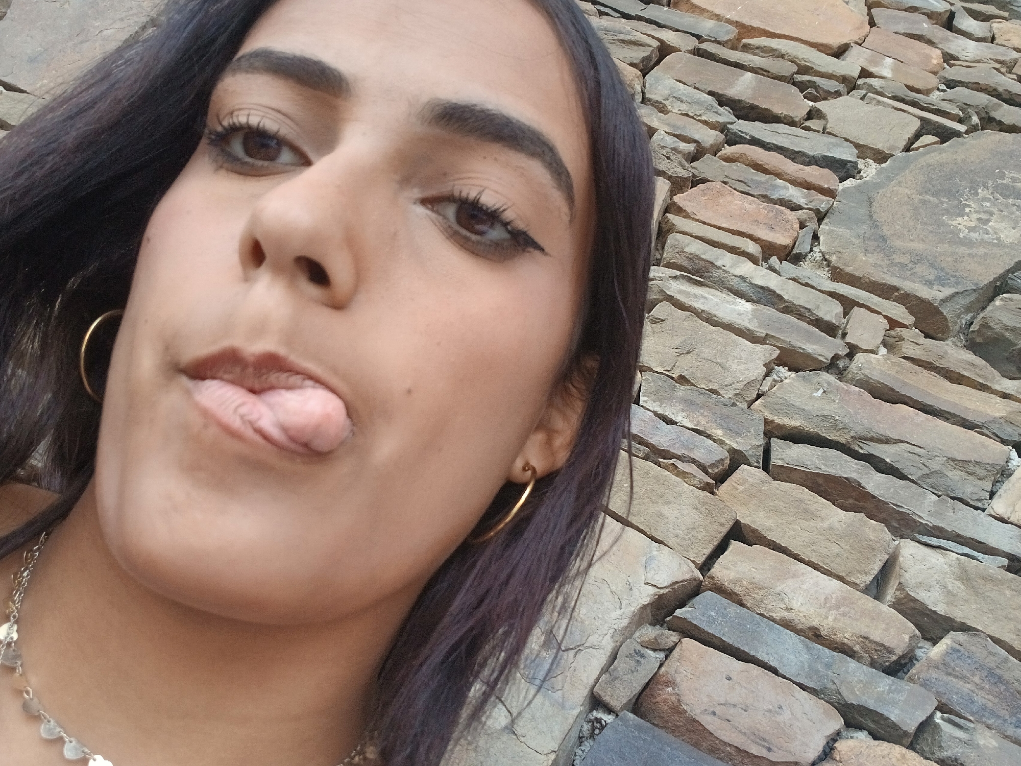 A selfie shot of a dark-haired woman against a stone wall. She has brown eyes, defined eyebrows, and eyeliner. She is sticking her tongue out. She is wearing gold hoop earrings and a delicate silver necklace. The background appears to be a wall constructed of stacked, flat stones of varying shades of brown and gray. The location is possibly near a stone wall in Juan Griego, Venezuela, based on the query.