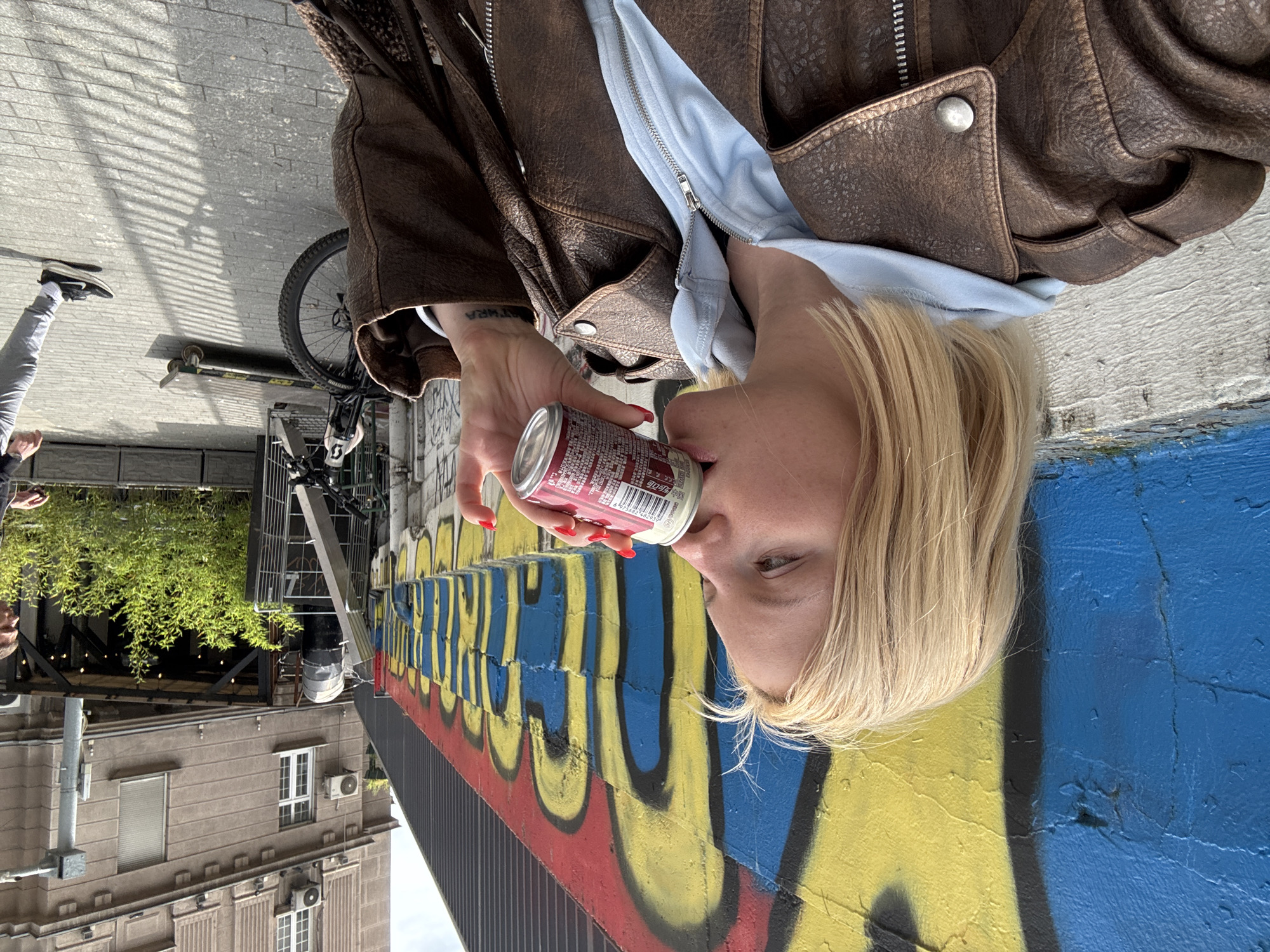 A blonde woman is drinking from a red and white can of soda while standing outdoors. She is wearing a brown leather jacket over a light blue hooded sweatshirt. Behind her is a colorful graffiti mural on a concrete wall, primarily featuring blue and yellow colors.
In the background, urban elements include a bicycle, a building with windows, greenery, and a sidewalk. Other people are partially visible on the left side of the image.
The location is Belgrade, Serbia.