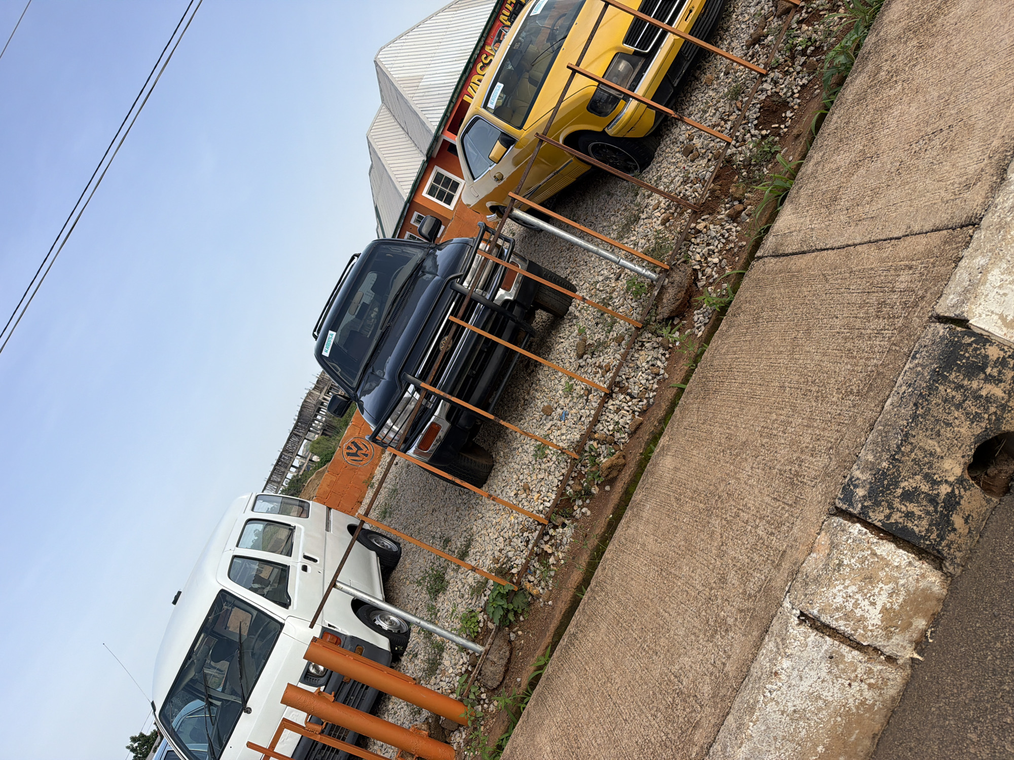 In Bukuru, Nigeria, a white van is parked behind an orange metal fence, next to a black SUV and a yellow sedan. The van has its headlights on and wipers on the windshield. The SUV has a spare tire mounted on the rear and a roof rack. The yellow sedan has a sticker on its windshield. The vehicles are parked on a gravel lot. In the background, there is a building with a white roof and orange walls. The sky is clear and blue.