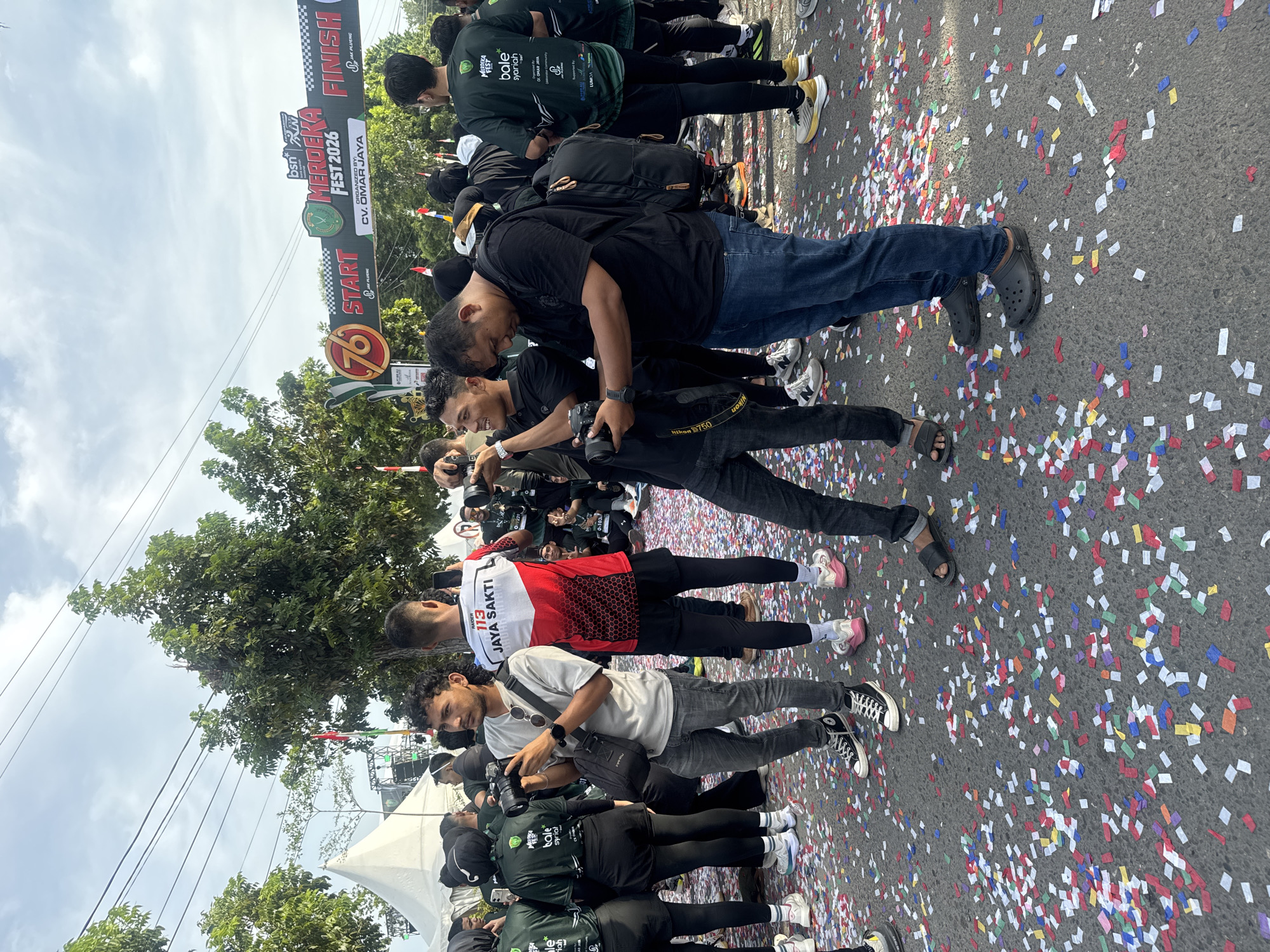 The image depicts an outdoor scene in Lhokseumawe, Indonesia, possibly near a finish line or starting point of an event, indicated by a banner that reads "MERDEKA FEST 2026 START" and "FINISH". The ground is covered with a significant amount of colorful confetti, suggesting a celebratory atmosphere.
Several individuals are present, many holding cameras, suggesting they are photographers or participants documenting an event. The people are diverse in age and gender, dressed in casual attire, athletic wear, and some in green t-shirts with event branding. One person in the foreground, wearing a gray t-shirt and jeans, is actively holding and using a camera. Another individual in a black shirt and blue jeans is looking down at their phone. A person in the background wears a red and white athletic jersey with the text "113 JAYA SAKTI".
Tents and structures are visible in the background, along with trees and overhead power lines. The sky is overcast with some visible clouds. The overall impression is of a lively event with a focus on photography and community gathering.