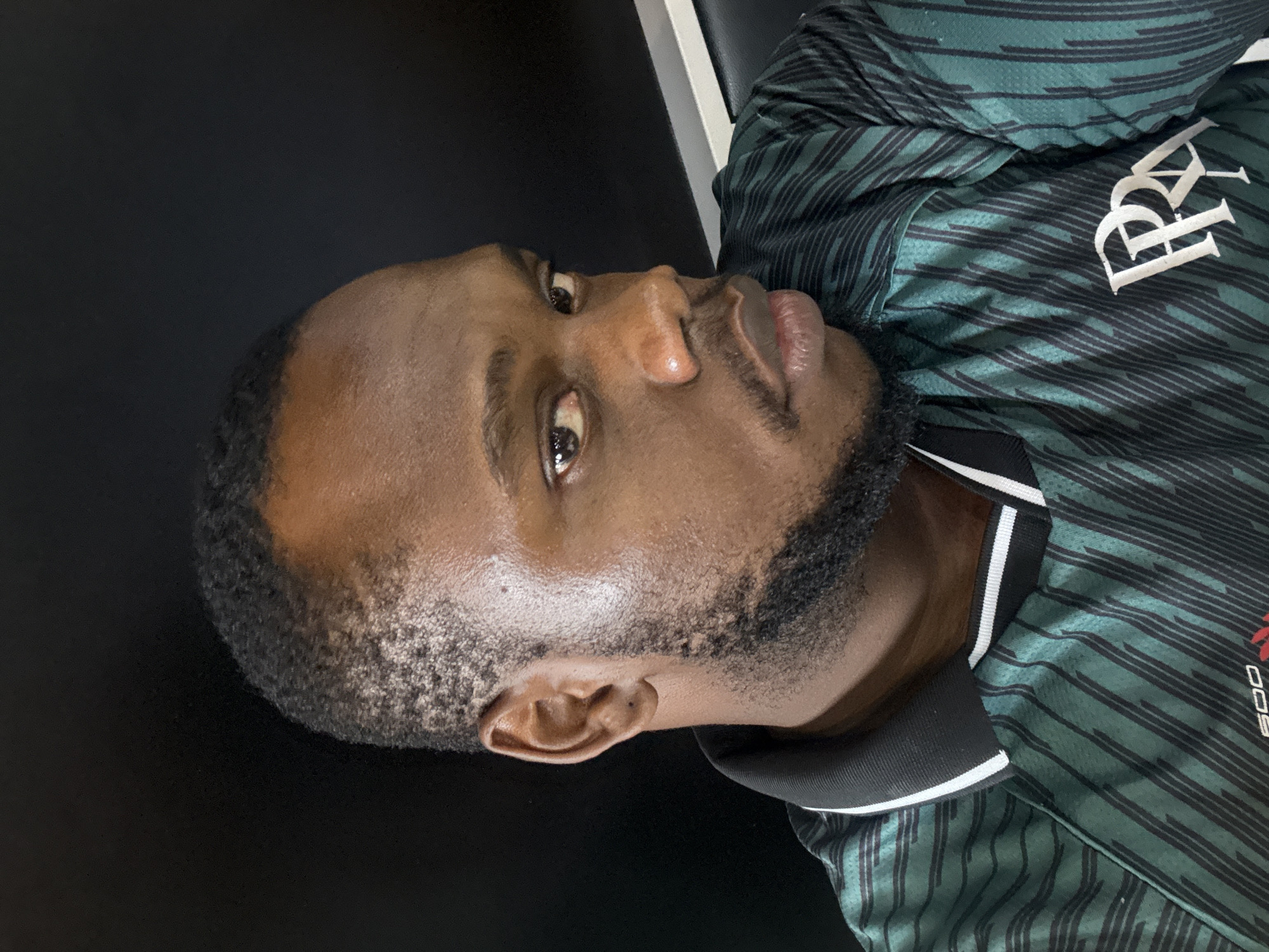 The image is a close-up shot of a dark-skinned man wearing a green and black striped collared shirt with white lettering. The man has short, dark hair and a beard. The background appears to be a dark, solid color. There is no information in the image to indicate that it was taken in Lugbe, Nigeria.