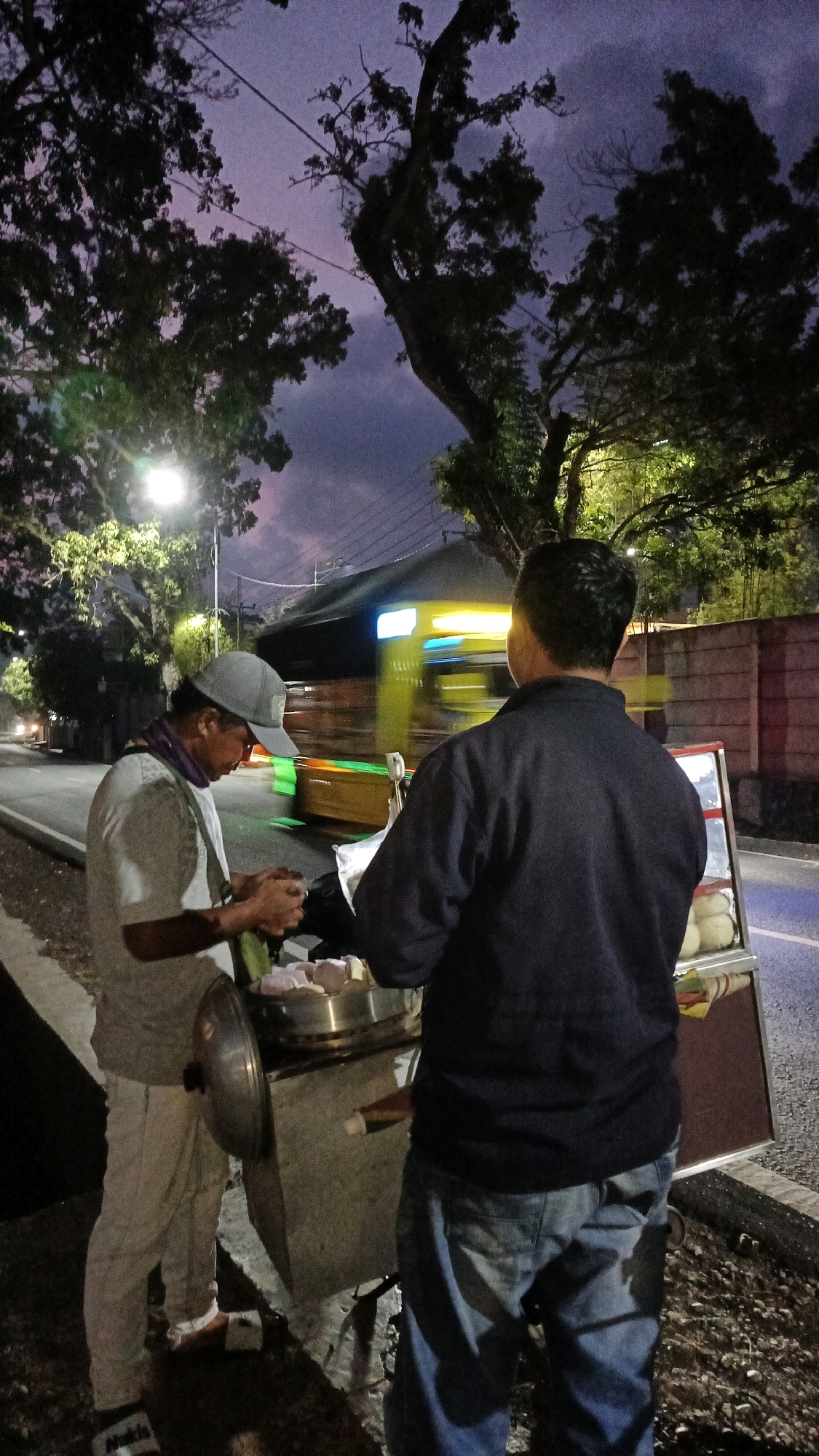 In Sukaresik, Indonesia, two men are standing on the side of a road at dusk. The man on the left, wearing a grey cap, a white shirt, and light-colored pants, is preparing food at a mobile cart. He is holding a piece of food in his hands. The man on the right, wearing a dark blue jacket and blue jeans, is standing behind the cart, looking at the food being prepared. The cart appears to be selling steamed buns or similar food items, with several buns visible on display. A blurred yellow and green vehicle is passing by on the road behind them, indicating a moderately busy street. The sky is a deep purple and pink, with silhouetted trees and a street lamp illuminating the scene. The ground is dark and damp, suggesting recent rain or humidity. A concrete wall is visible in the background to the right.