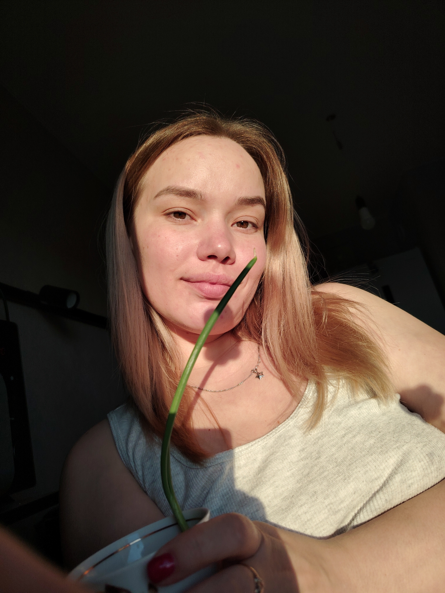 A young woman with blonde hair and fair skin poses for a selfie indoors. She is wearing a grey tank top and a delicate necklace with a star pendant. She holds a small cup or saucer in her hand, from which a single, green plant stem extends upward, partly obscuring her face. Her expression is neutral, with a slight smile. The background is split into a dark, shadowy area and a brightly lit area, suggesting the presence of strong sunlight coming from a window or other light source. The location is possibly in the city of Марковский, Russia.