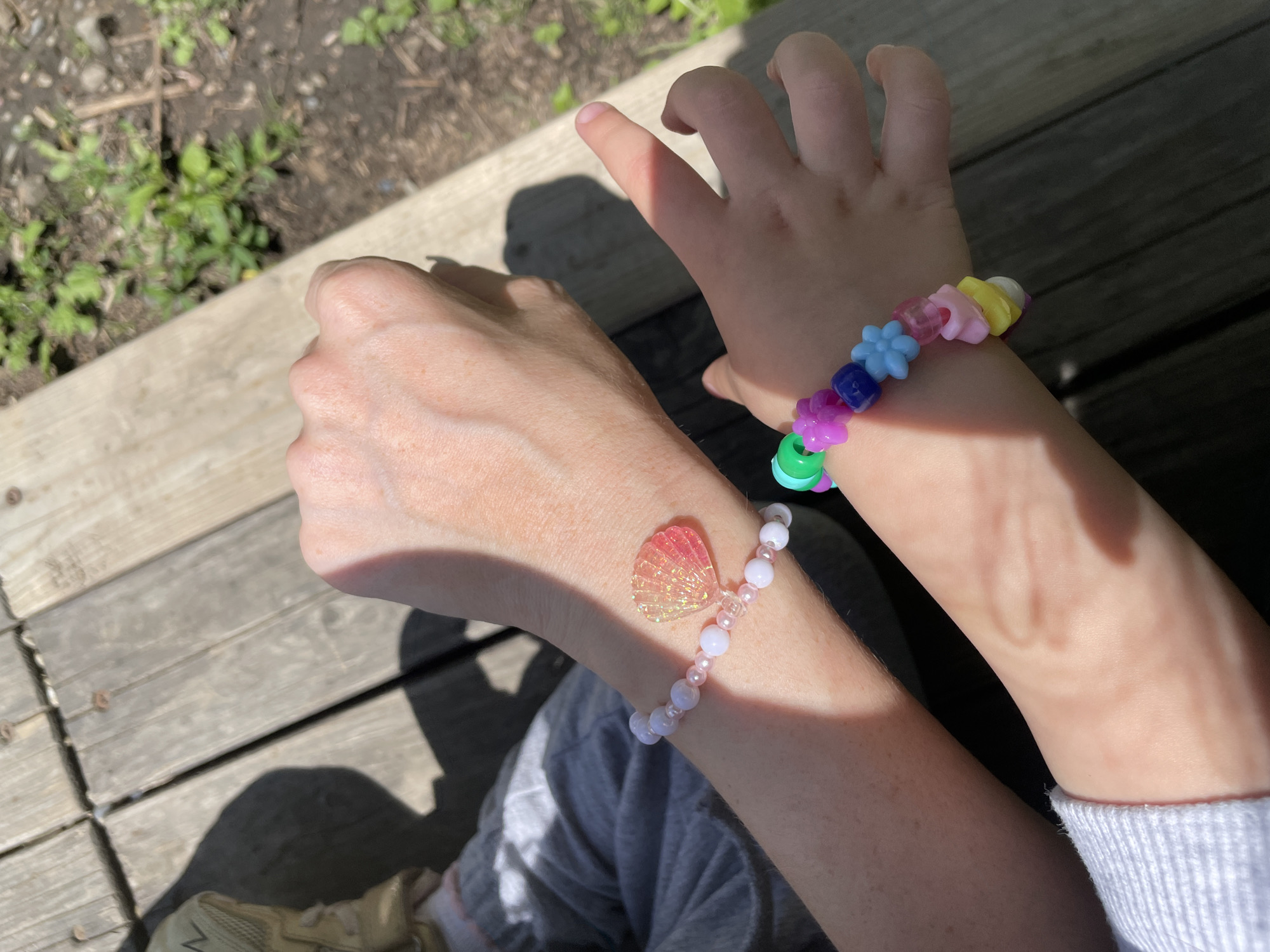 The image shows two arms, likely a child's and an adult's, resting on a wooden surface that appears to be a deck or platform. The child's arm, closer to the top right, has a colorful plastic bead bracelet with flower-shaped beads. The adult's arm, on the left, wears a bracelet with light pink beads and a seashell charm. There is a shadow cast on the wood. The background shows glimpses of green foliage, possibly grass or plants. Part of a shoe is visible in the lower left corner. The location is not identifiable as Vladivostok, Russia, or any other specific place based on the visual information provided.