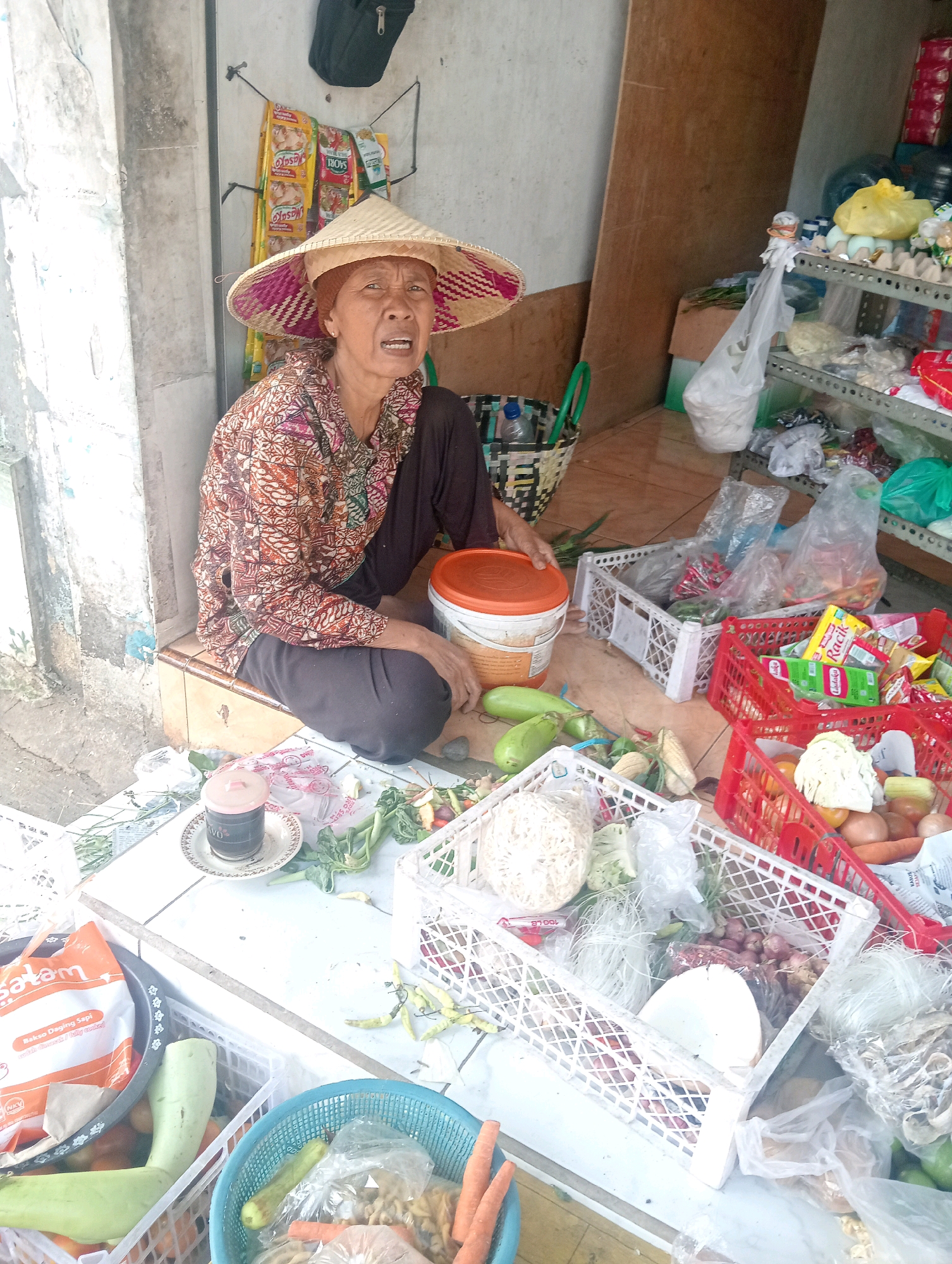 A street vendor, likely an elderly woman wearing a conical straw hat and a patterned shirt, sits on a low concrete ledge in Bojonegoro, Indonesia. She is surrounded by baskets and containers filled with fresh produce, including vegetables like gourds, chilies, onions, cabbage, and carrots. Packaged goods and other items are visible on shelves in the background, suggesting a small roadside stall. The vendor appears to be engaged in her work, with a cup of dark liquid, possibly coffee, placed on a small table beside her. The scene depicts a typical urban market environment in Indonesia.