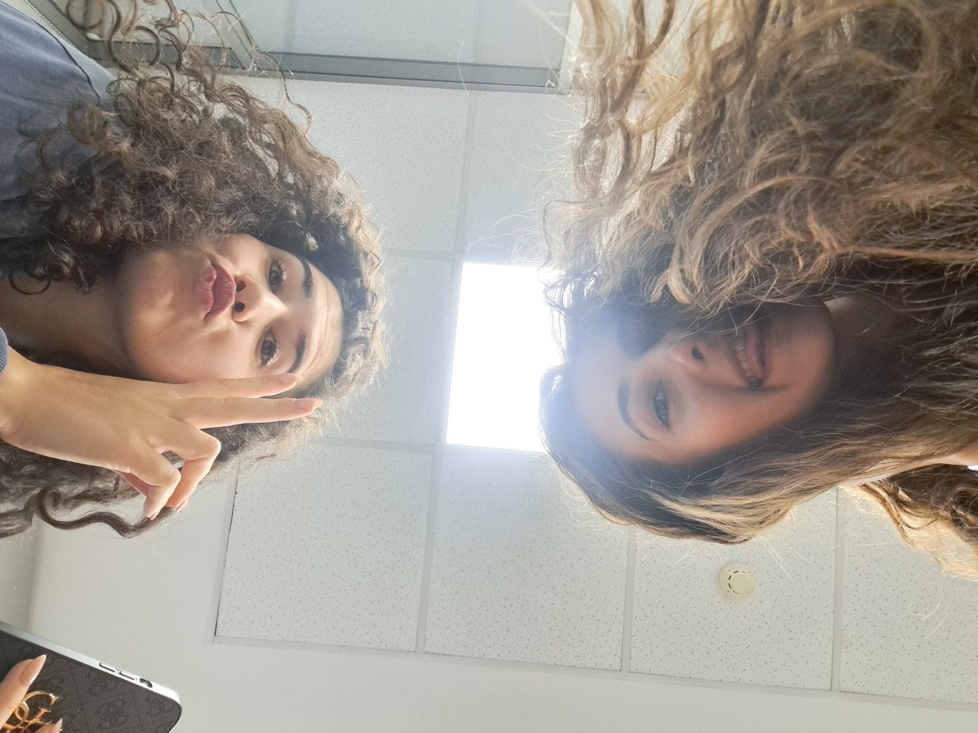 This low-angle image features two young women looking down into the camera, likely posing for a selfie. The setting is indoors, characterized by white acoustic ceiling tiles, a prominent bright rectangular light fixture in the center background, and a circular smoke detector on the ceiling near the right woman.
The woman on the left has dark, curly hair and is wearing a blue top. She makes a playful pouting expression with her lips and forms a peace sign with her right hand. She holds a phone with a brown case that displays a gold "G" logo, indicative of the Guess brand. Her eyes are open and directed towards the camera.
The woman on the right has lighter, wavy hair. She is smiling broadly, revealing her teeth, and her eyes are also open and looking down. A bright glare emanates from the overhead light fixture, particularly affecting the right side of the image and the woman's hair. Both individuals exhibit cheerful and relaxed expressions. No other discernible text is visible in the frame.