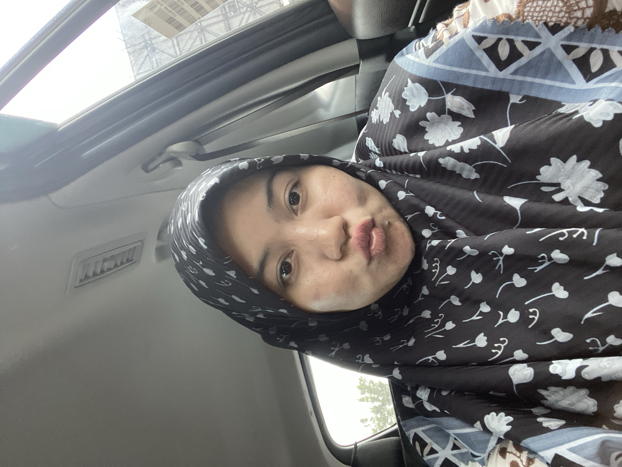A woman wearing a black and white floral hijab and a patterned shirt is taking a selfie inside a car. She is puckering her lips and looking at the camera. The interior of the car, including the ceiling, air vent, and window, is visible. Outside the car window, trees and a distant structure are visible. The scene is lit by natural light. The location is identified as Banjarbaru, Indonesia.