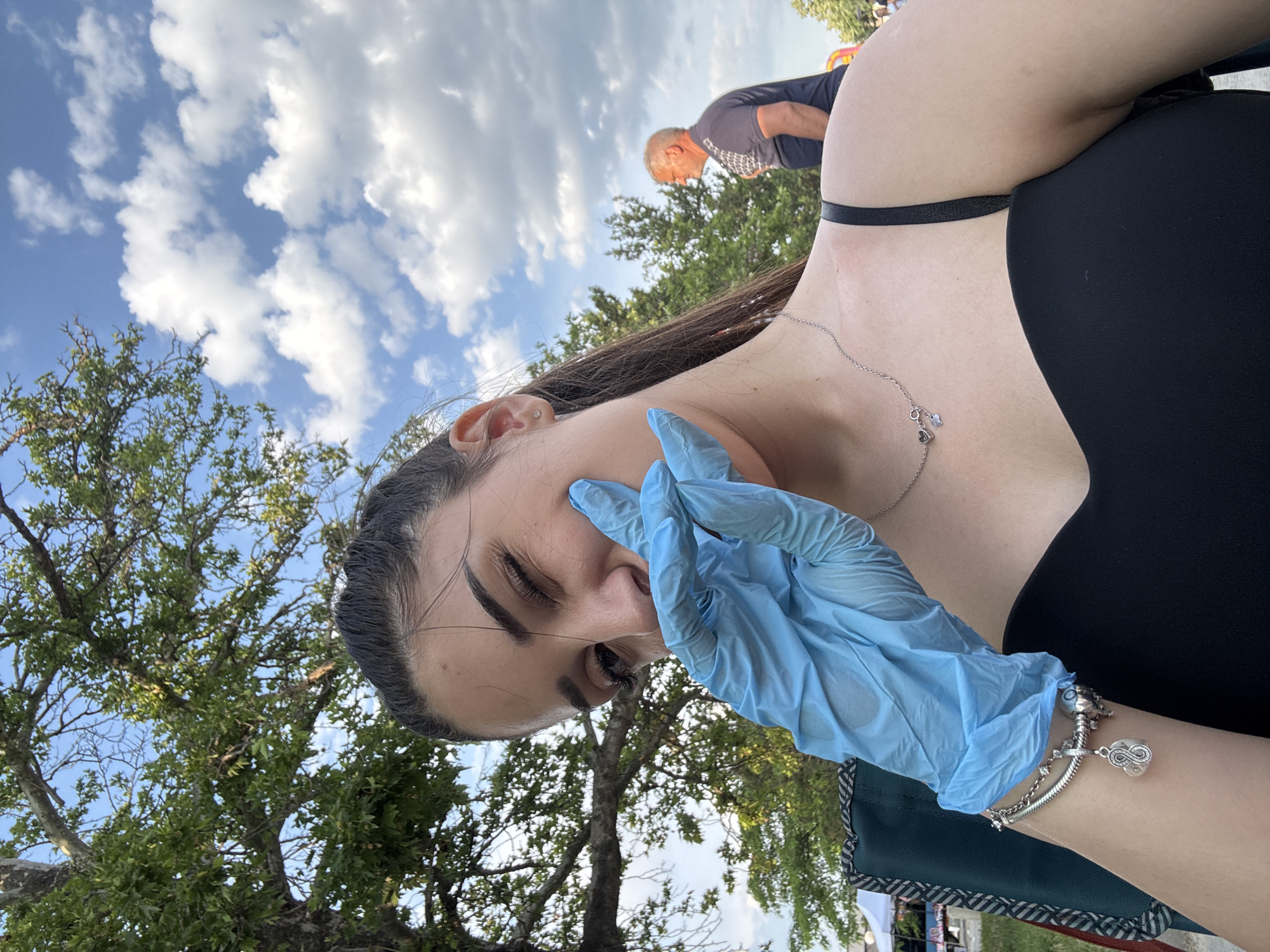 An outdoor close-up shot captured in the city of Kostievo, Bulgaria, features a young female subject prominently in the foreground. She has dark hair styled back and is winking with her left eye. Her right hand, covered by a light blue disposable glove, is placed against her cheek, partially obscuring her mouth. She is wearing a black strapless top or dress, a silver necklace with two small charms, and a silver charm bracelet on her left wrist. A segment of a green fabric chair is visible in the lower left. The background consists of a clear blue sky with scattered white cumulus clouds and abundant green tree foliage, including a large deciduous tree to the left. In the mid-ground on the right, an older male with grey hair, wearing a dark blue polo shirt, is partially visible from the chest up, facing away from the camera. A white tent or canopy structure is faintly discernible in the far left background.