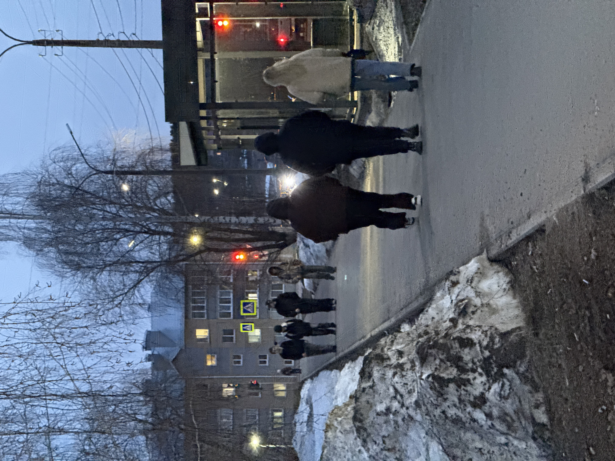 The image depicts an urban scene in what appears to be Votkinsk, Russia, based on the architecture. A group of people are walking along a paved sidewalk. The foreground features melting snow along the sidewalk edge.
In the background, there is a multi-story residential building, some windows are illuminated. A pedestrian crossing sign is visible.
A bus stop is situated nearby, with people waiting inside. Overhead power lines and street lights are present. The sky is overcast.
