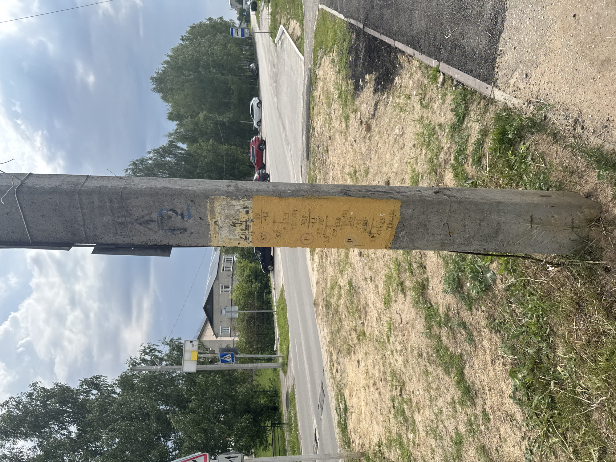 The image depicts a concrete utility pole with a yellow sticker affixed to it, covered in handwritten Russian text and numbers. The pole is located outdoors, near a paved road and a pedestrian crossing. In the background, there are trees, buildings, and a cloudy sky. The overall scene suggests an urban or suburban environment in the village of Krasnye Baki, Russia. The sticker on the pole appears to be a form of signage or informational display, though its exact purpose is unclear from the image.